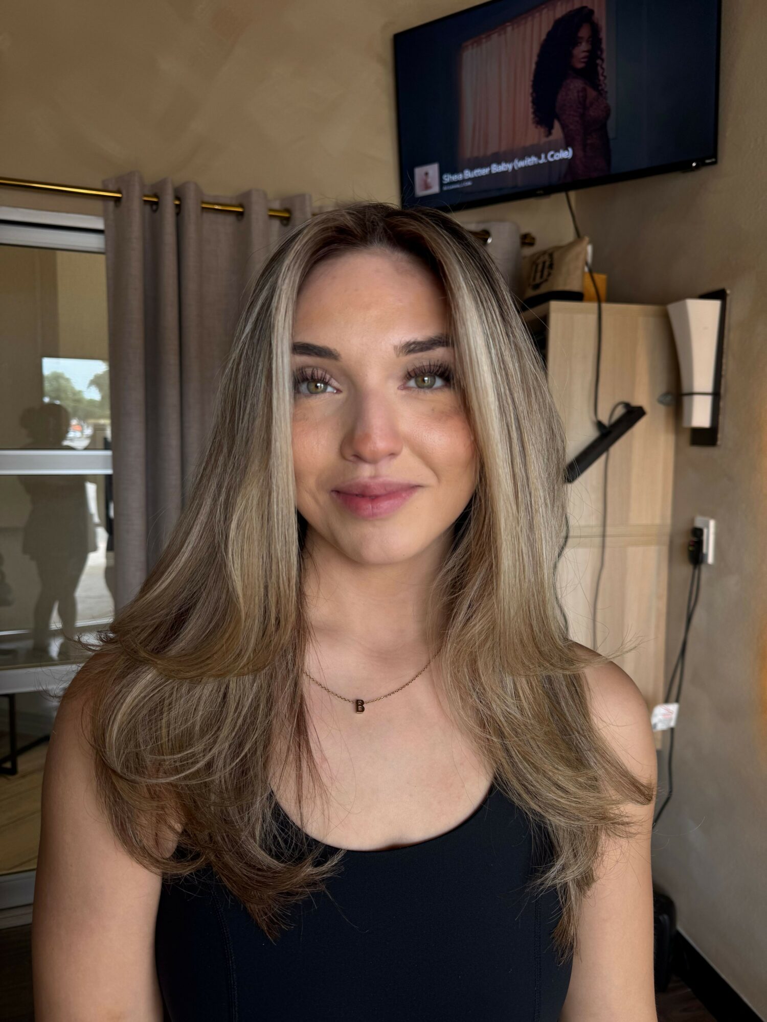 Young woman with long, wavy blonde hair and a black top, smiling indoors with a TV and window in background.