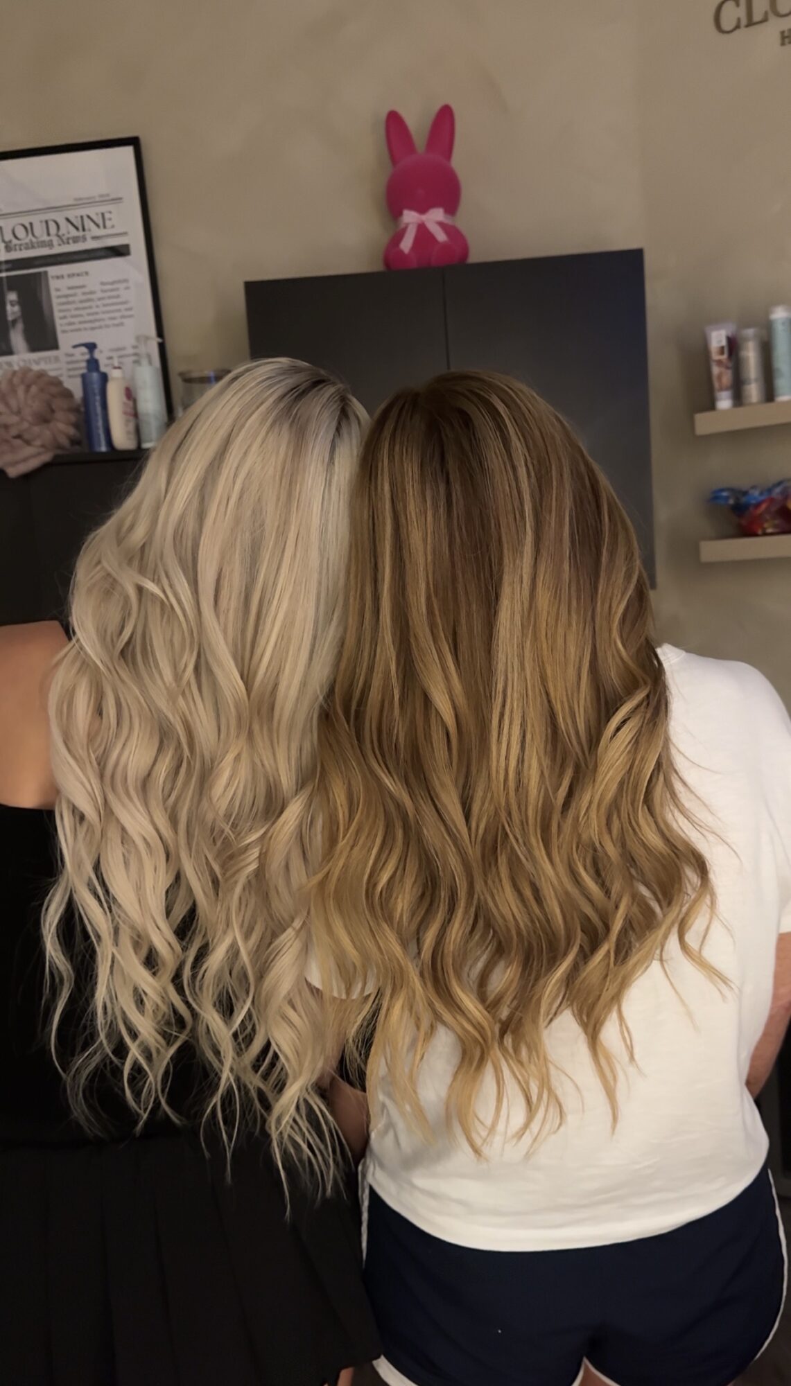 Two women with wavy hair, one blonde and one brunette, standing in a room with shelves and a pink bunny decoration.
