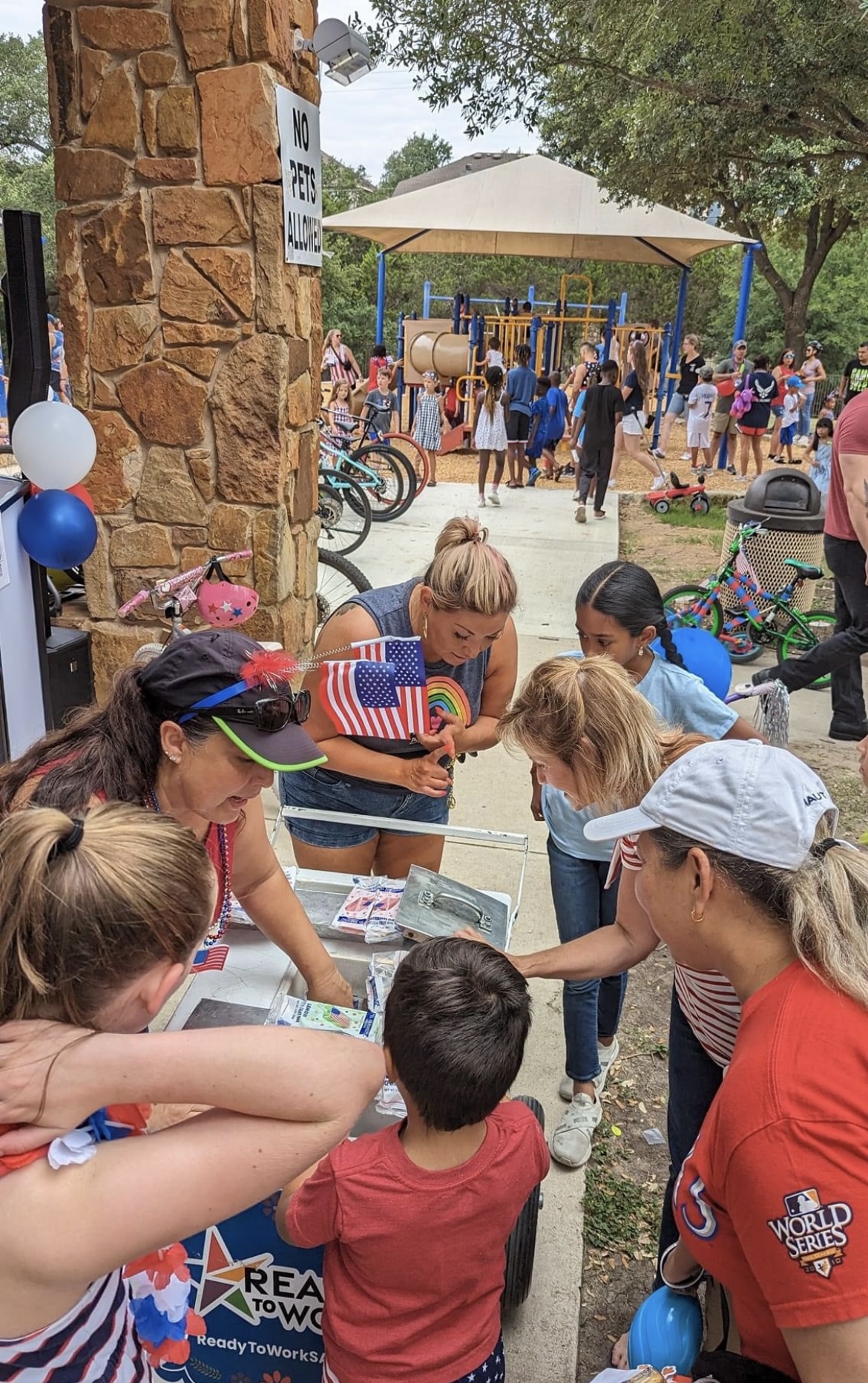 Children and adults gather around a table at an outdoor event, with a playground and crowd in the background.