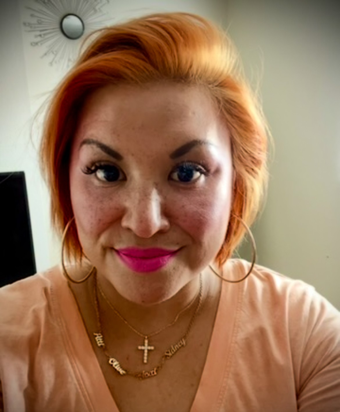 Close-up of a woman with orange hair, wearing pink lipstick, gold jewelry, and a light-colored top, smiling.