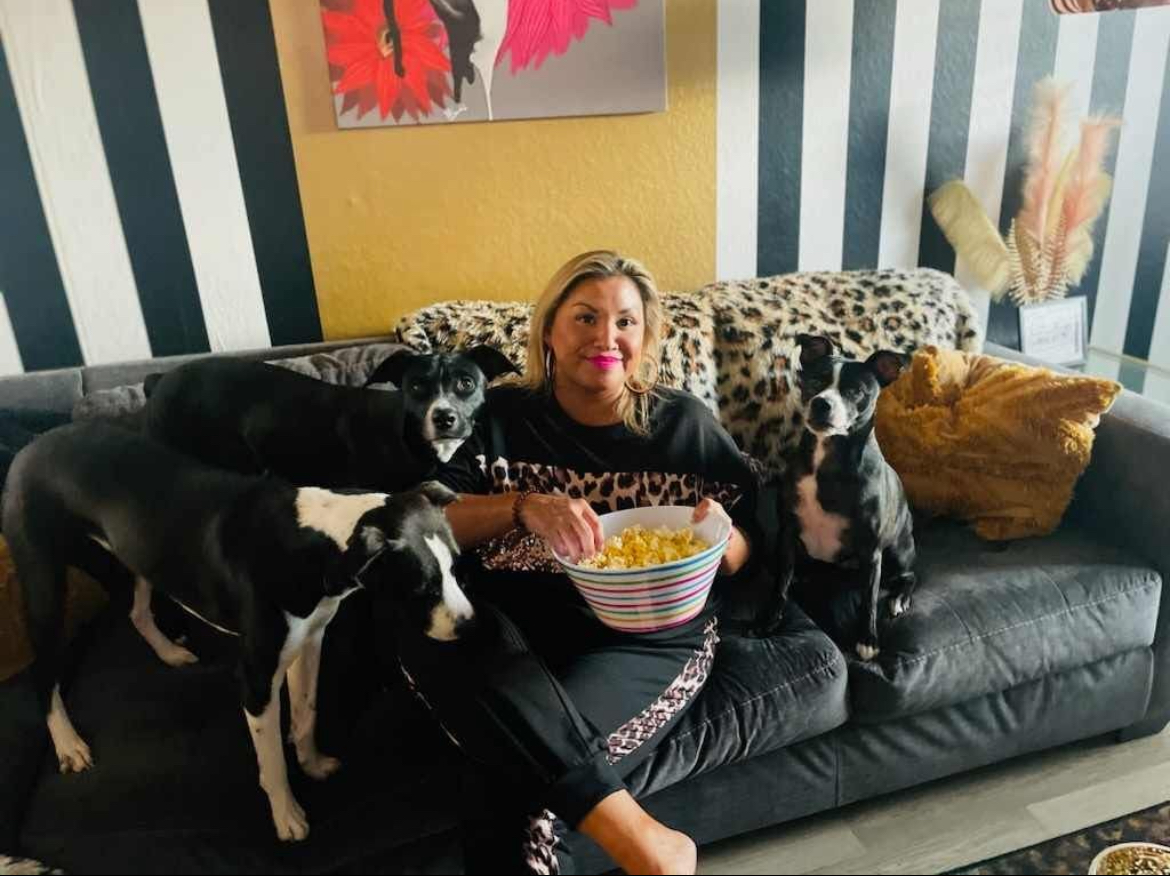 Woman sitting on a black sofa with five dogs around her, holding a bowl of popcorn, in a colorful room.