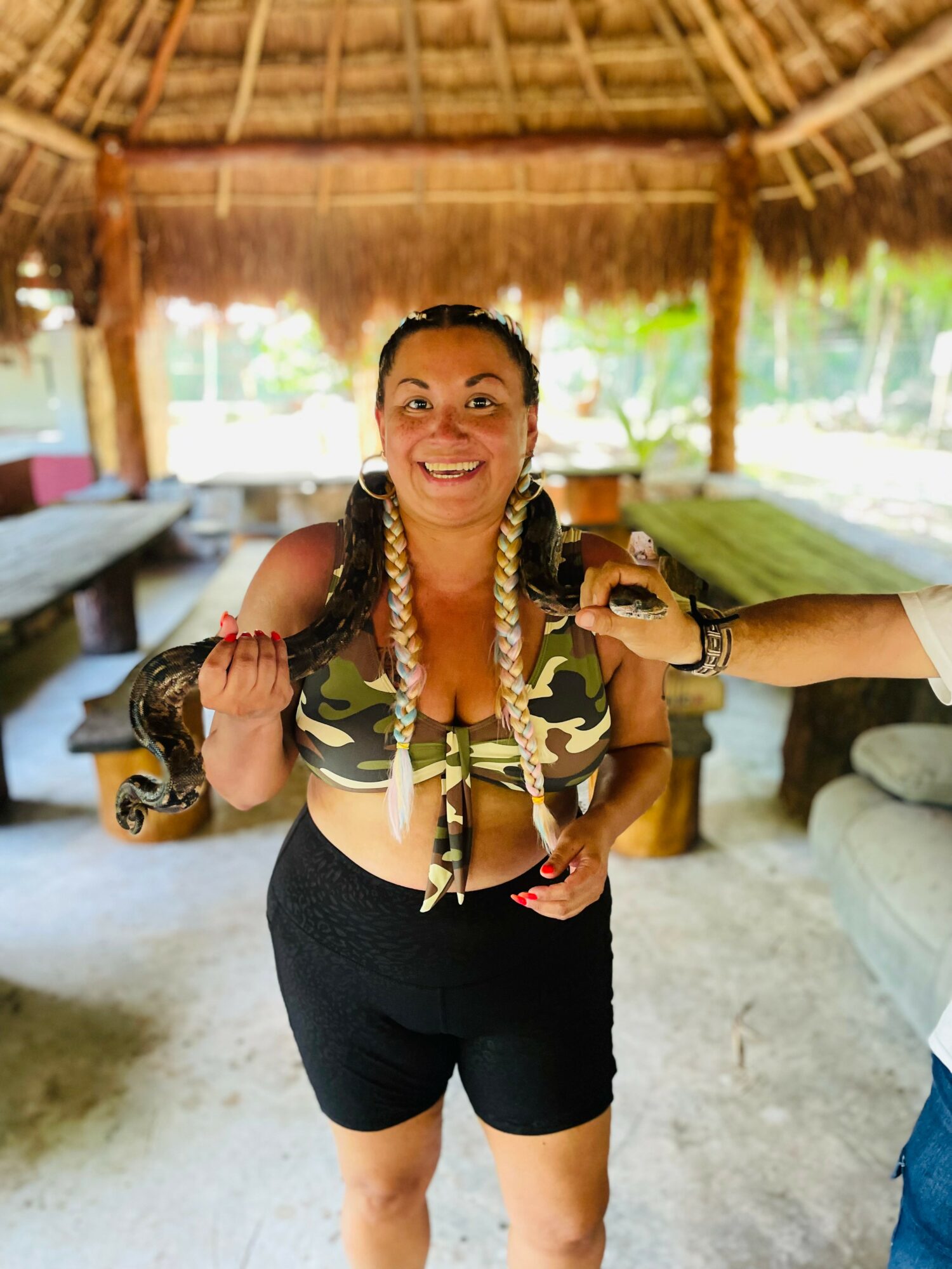 Woman smiling holding a snake, in a tropical hut setting with seating and greenery in background.