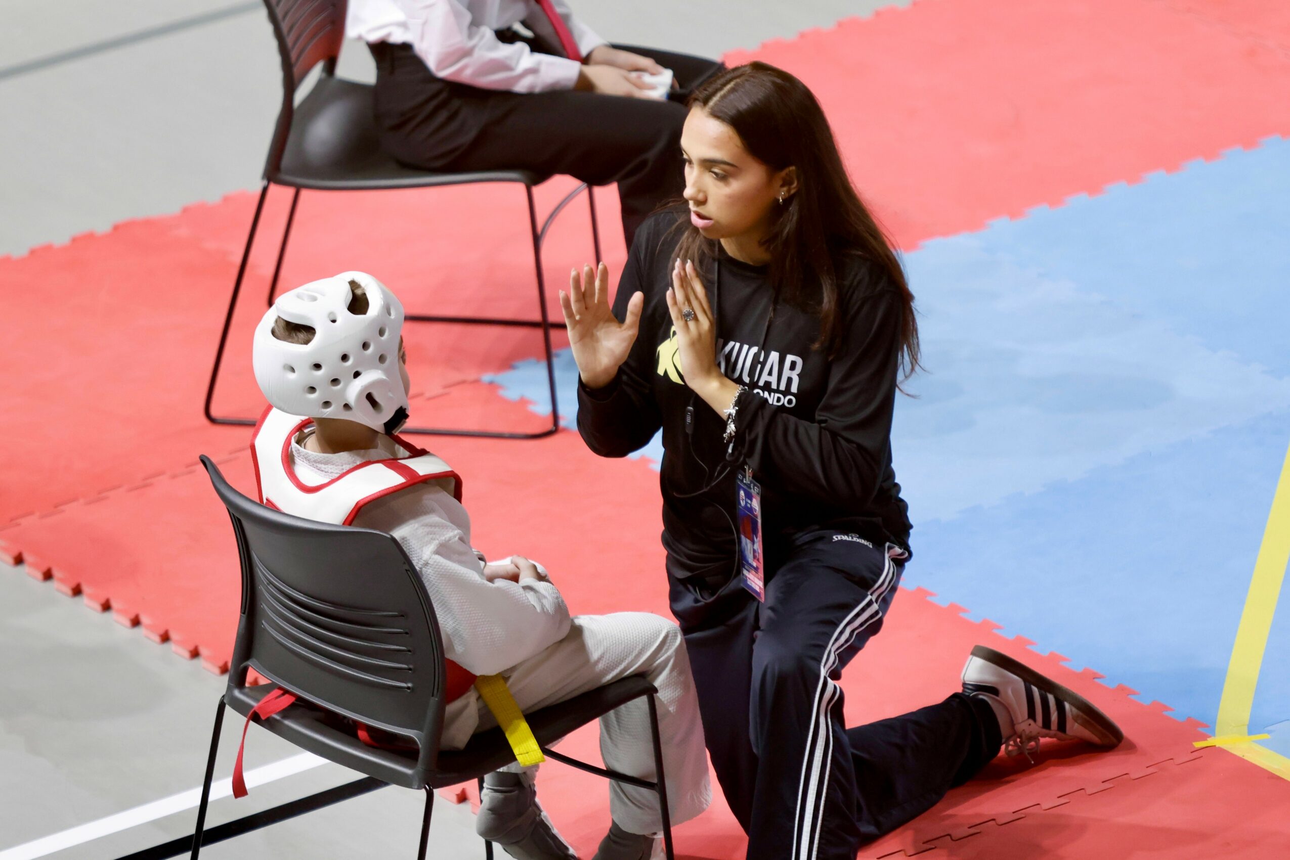 A woman kneeling on one knee, instructing a person in protective gear sitting on a chair, in an indoor sports area.