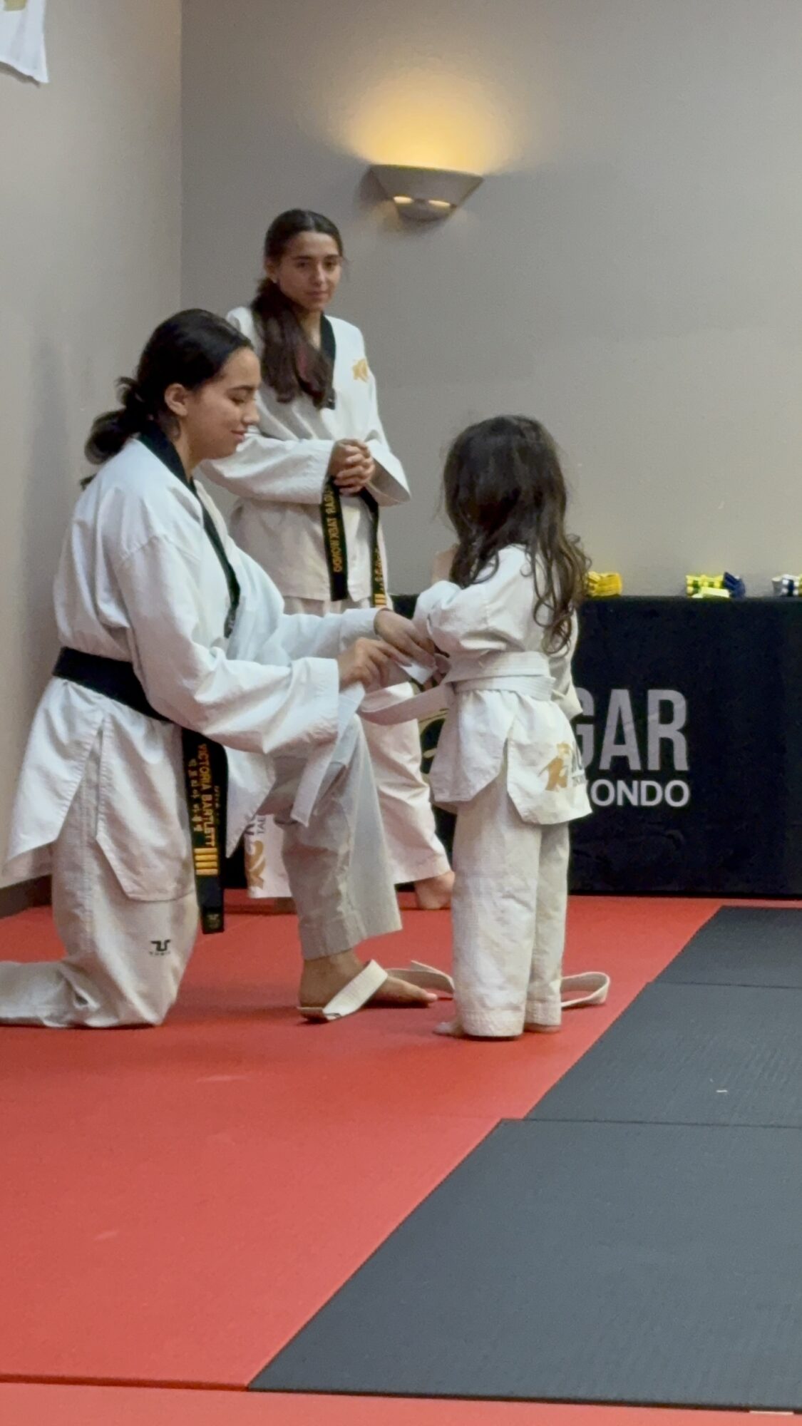 Two women in martial arts uniforms assist a young girl in a white martial arts uniform on a red mat, with a table in the background.