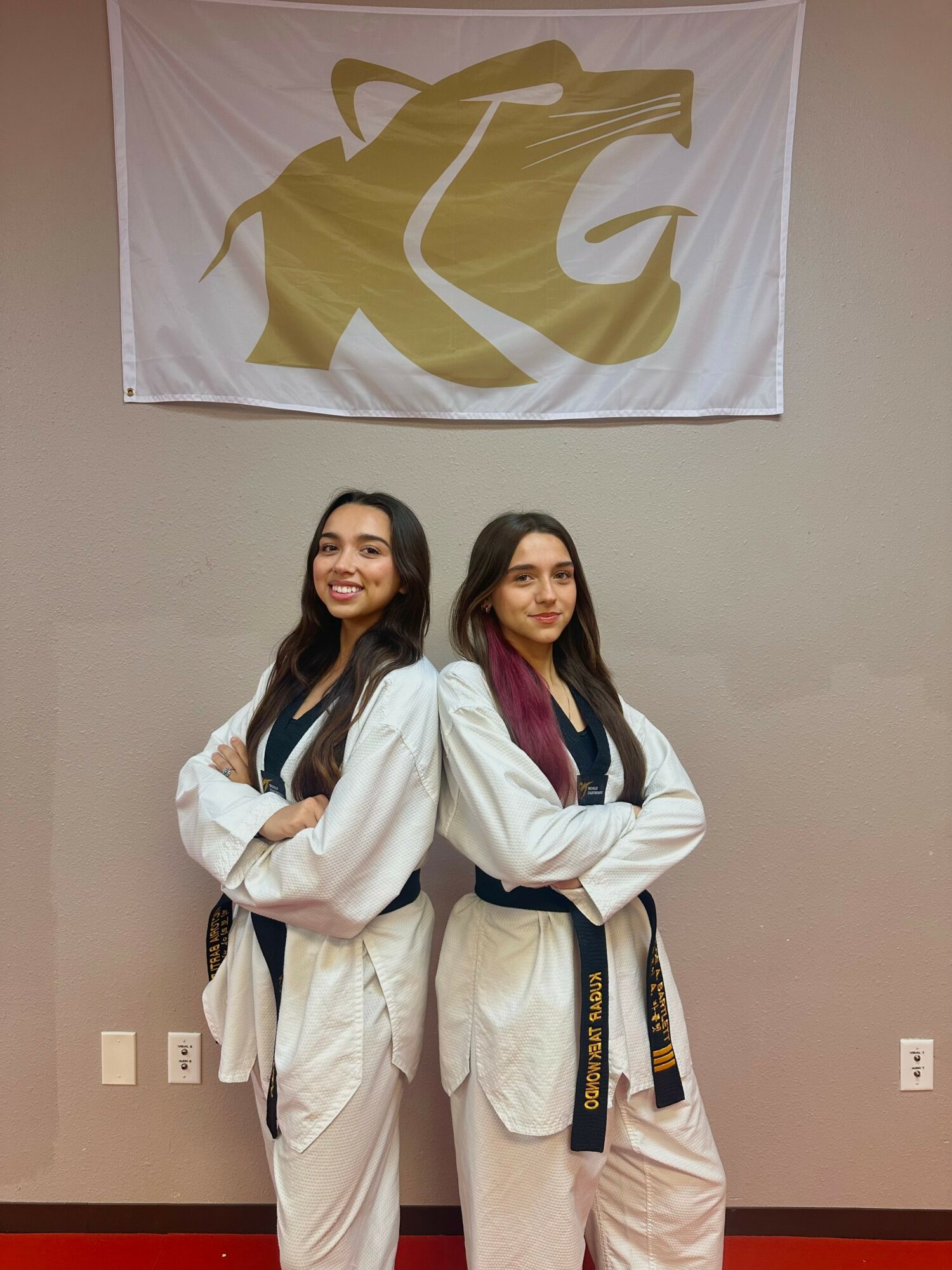 Two young women in martial arts uniforms stand side by side, smiling, with a large logo banner above them on the wall.