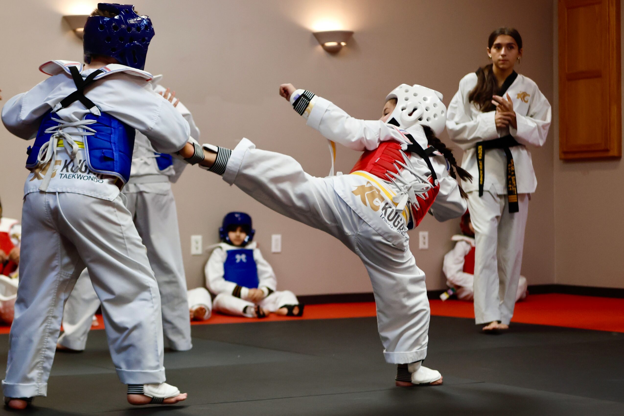 Martial arts students practicing kicks and techniques in a dojo, with a person observing in the background.
