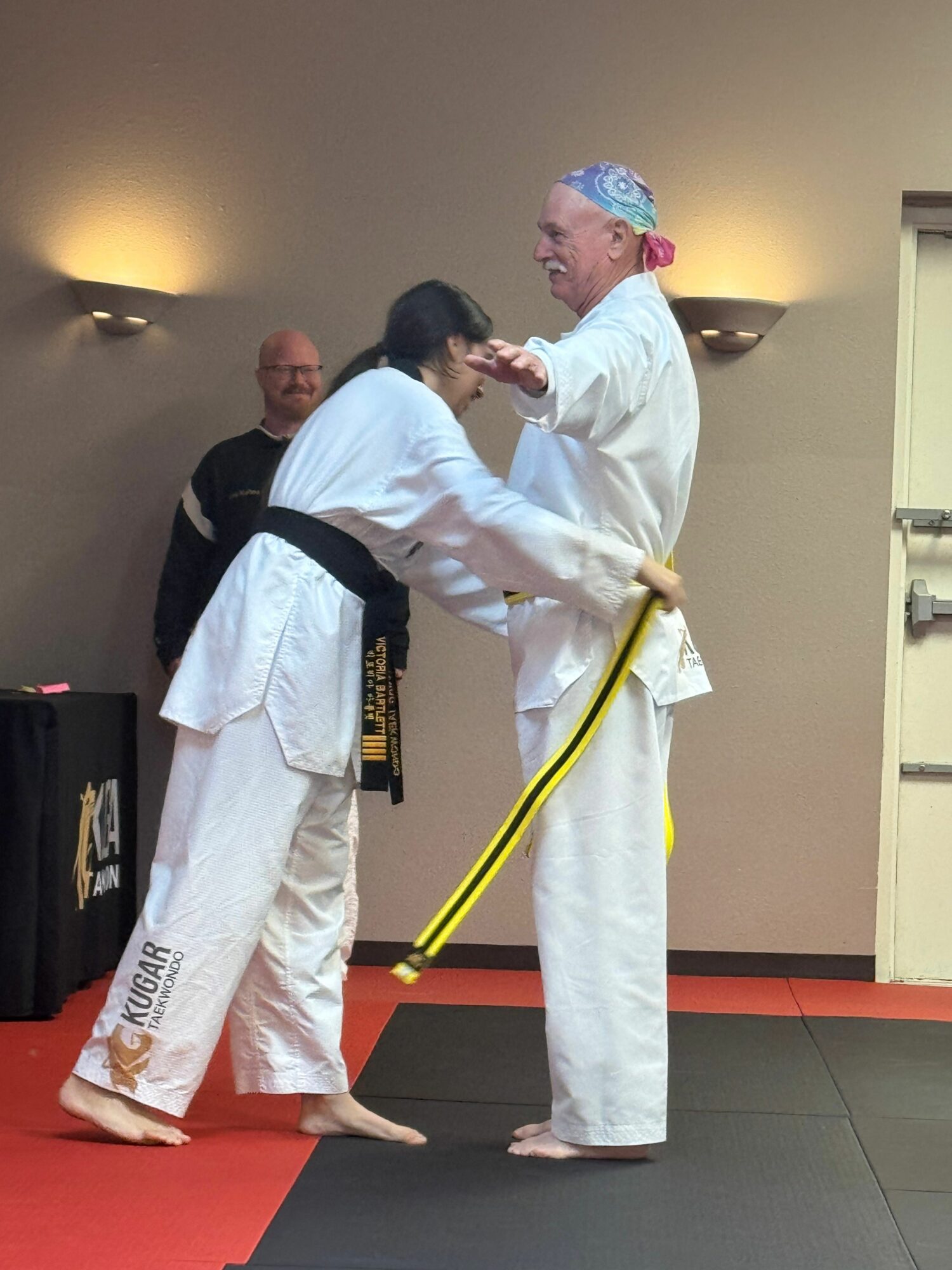 Two martial artists in white uniforms exchange a handshake or award, with a third person observing in the background.