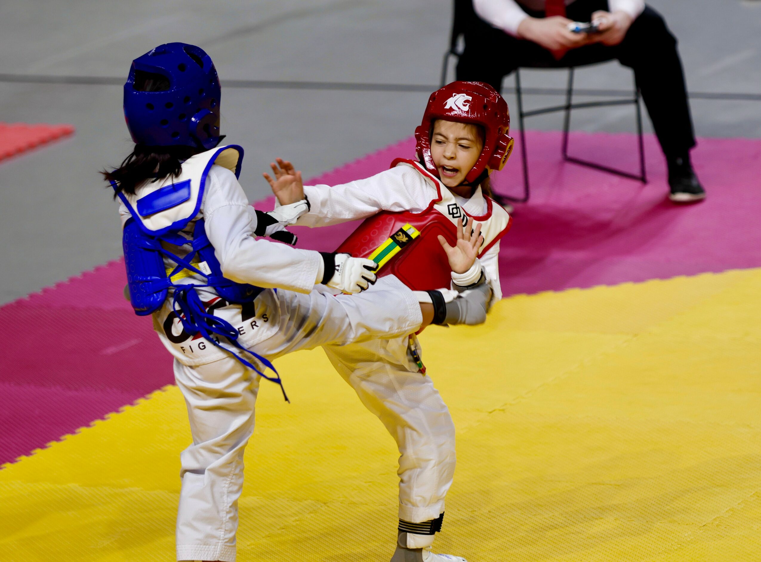 Two children in protective gear sparring on a yellow mat, one with a red helmet and the other with a blue helmet.