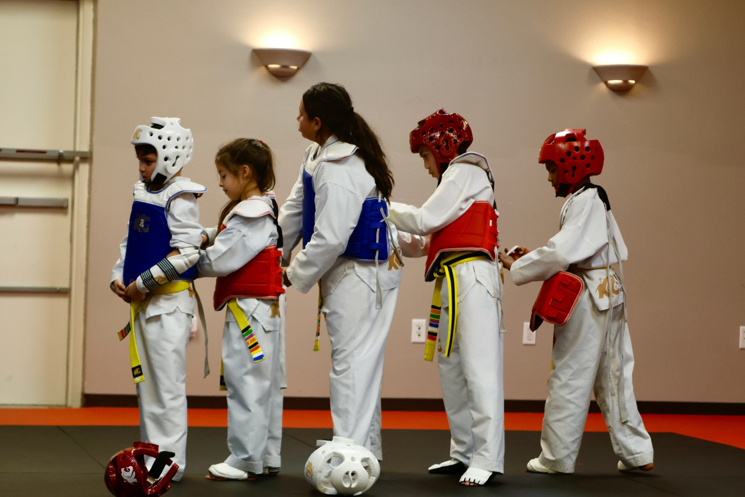 Group of children in taekwondo uniforms and protective gear standing in line indoors, with a soccer ball on the floor.