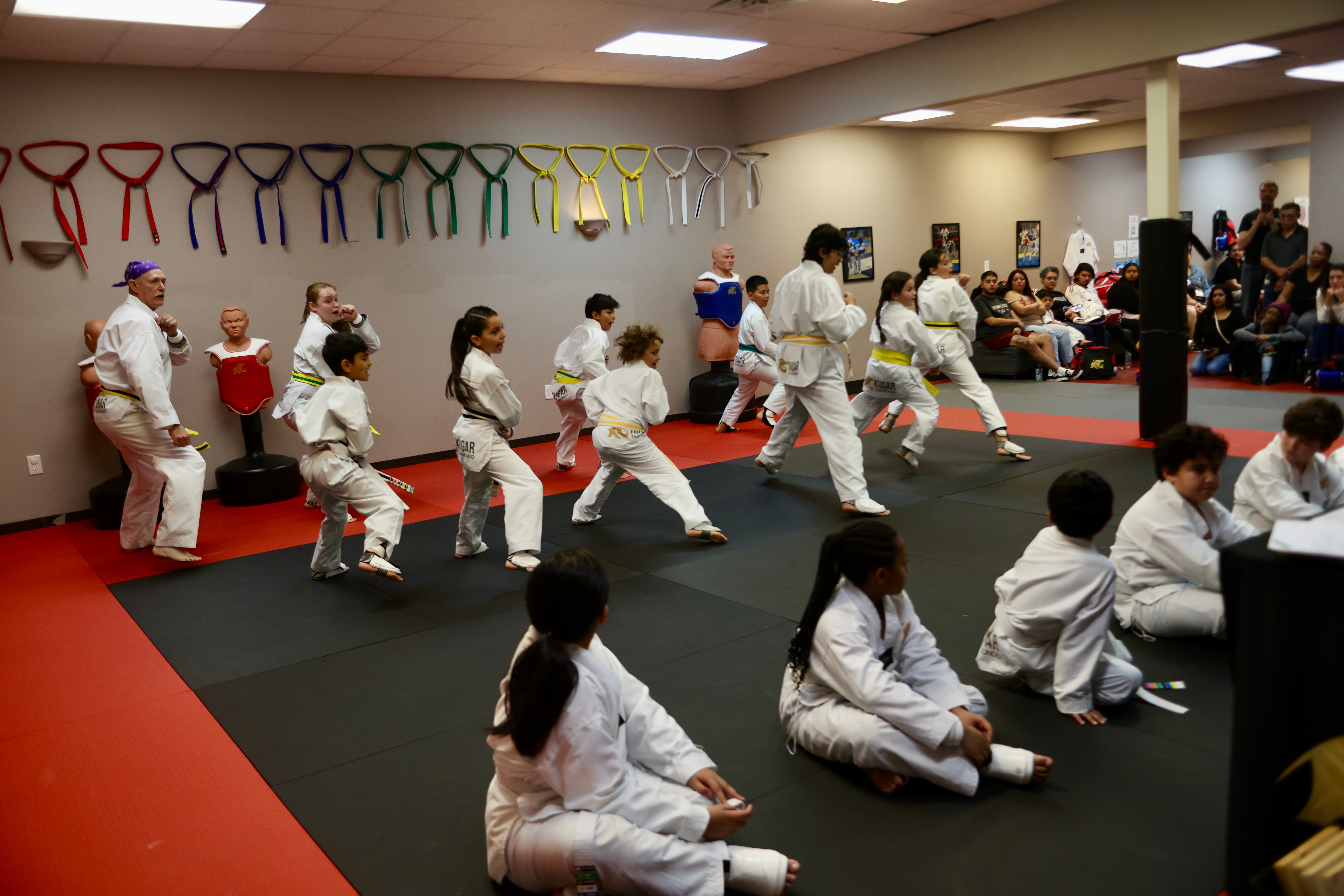 Children in martial arts uniforms perform synchronized moves in a dojo with colorful belts hanging on the wall.