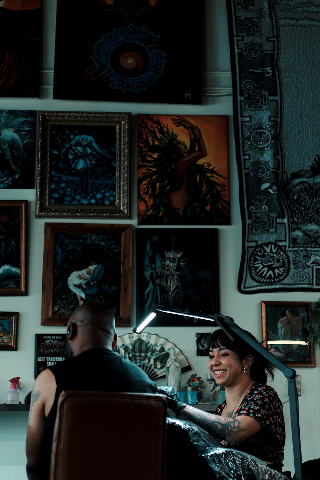 Two women sit in a room decorated with framed artwork and a tapestry, one smiling and the other with her back to the camera.