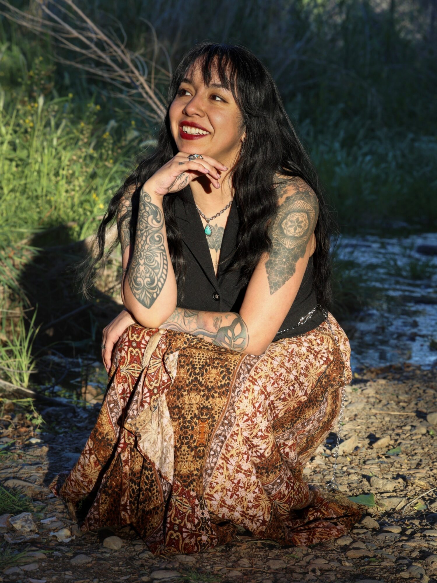 Woman with long dark hair, tattoos on arms, smiling outdoors near water and greenery, wearing a black top and patterned skirt.