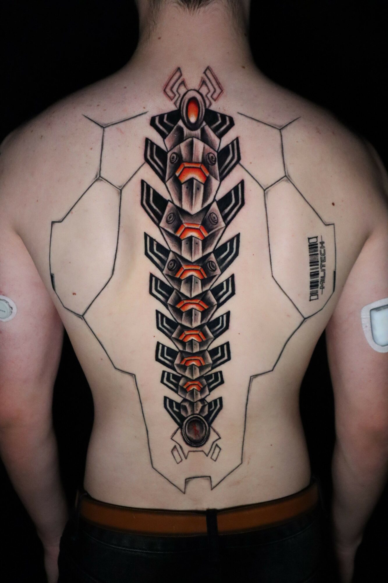 Back tattoo of a geometric, robotic insect with hexagonal patterns and orange accents, extending down the spine.