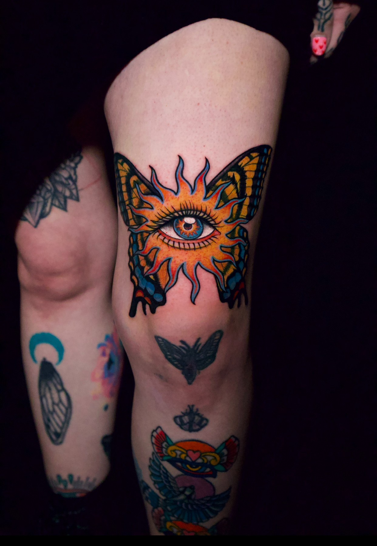 Tattoo of an eye with butterfly wings on a person's thigh, surrounded by other tattoos, against a dark background.
