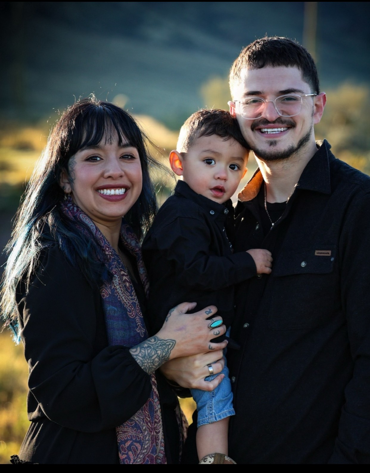 Three people outdoors, a woman, a man, and a child, smiling and posing together with a natural background.