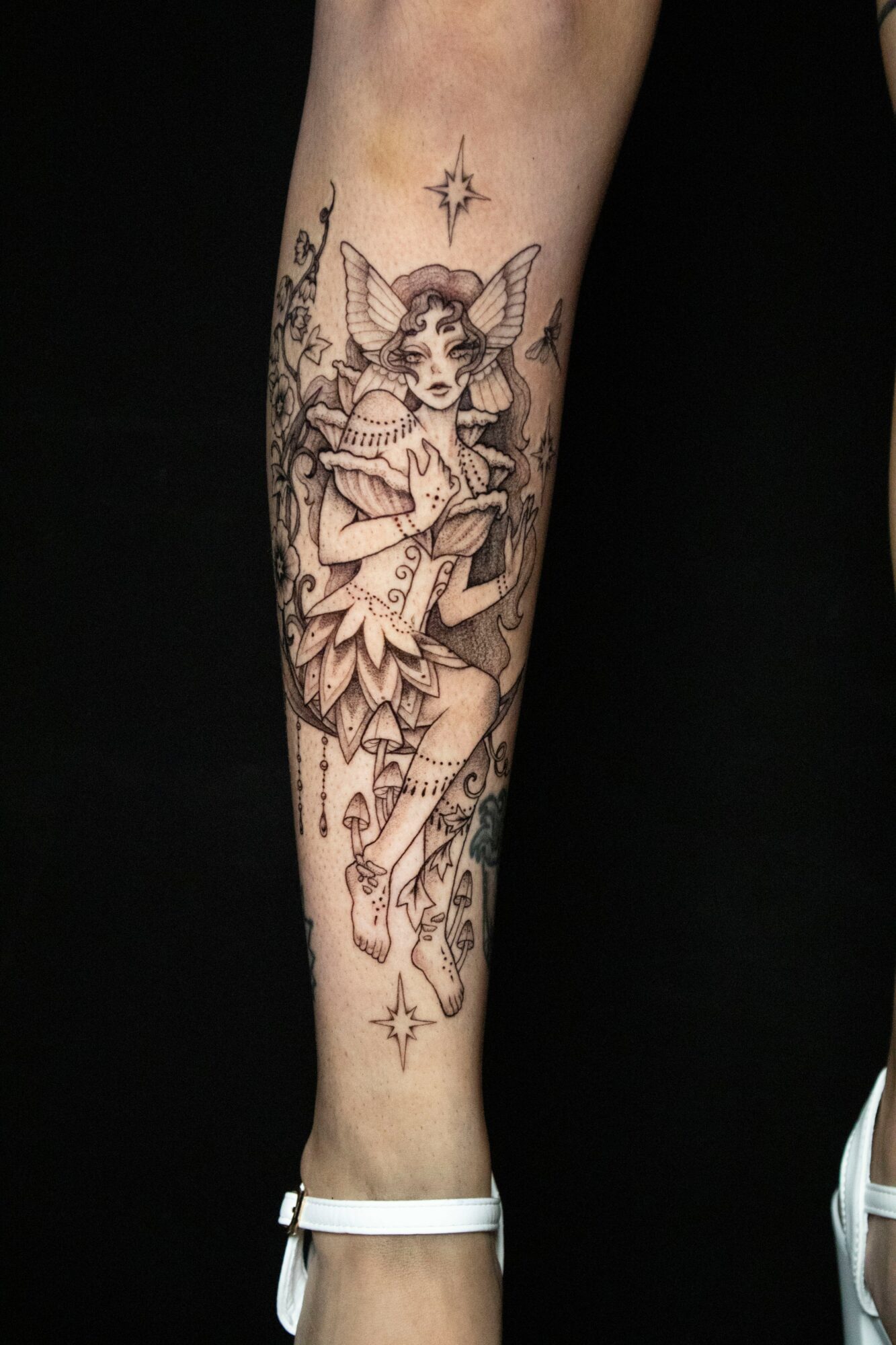 Black and gray tattoo of a fairy with wings, sitting on a branch, surrounded by stars and decorative elements, on a person's leg.