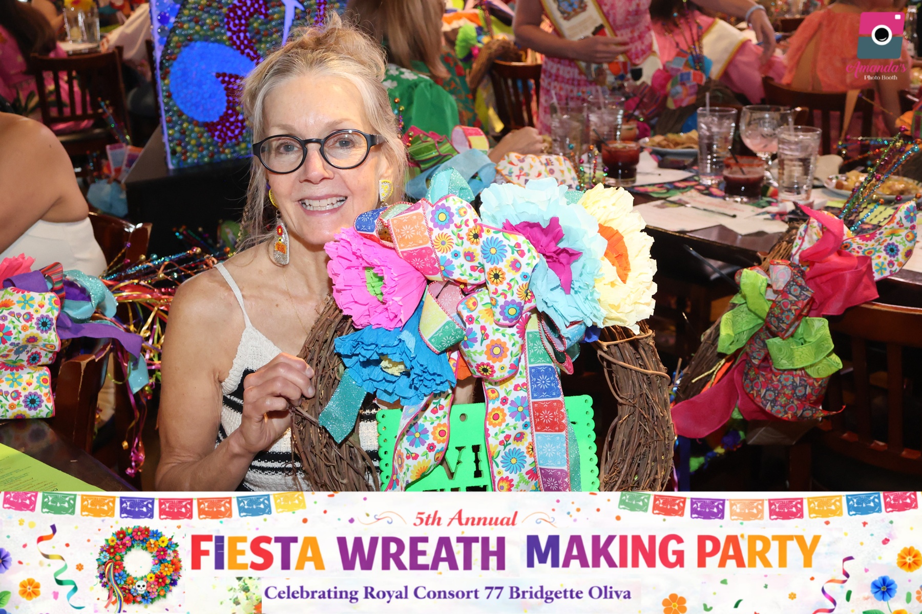 Elderly woman with glasses holding colorful fabric wreath at party, surrounded by people and tables with gifts and decorations.