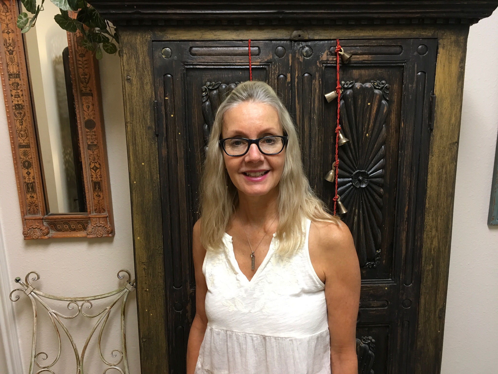 Smiling woman with glasses and long blonde hair standing in front of a dark wooden cabinet, wearing a white sleeveless top.