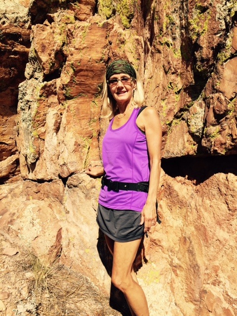 Woman in purple tank top and gray shorts standing outdoors against a rocky background, smiling, wearing a headband.
