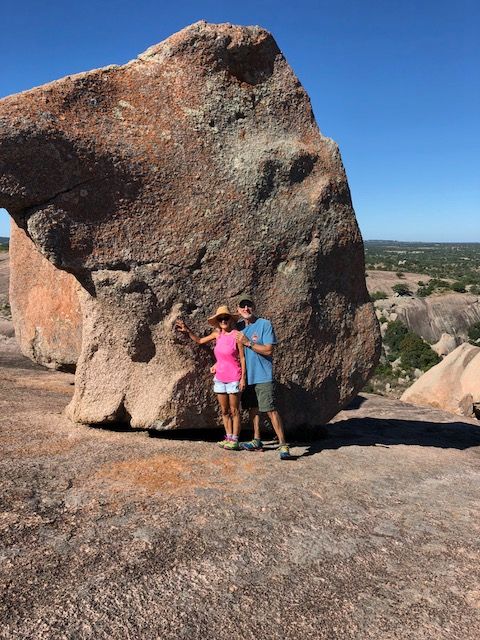 Two people stand next to a large rock formation in a desert landscape with a clear blue sky.