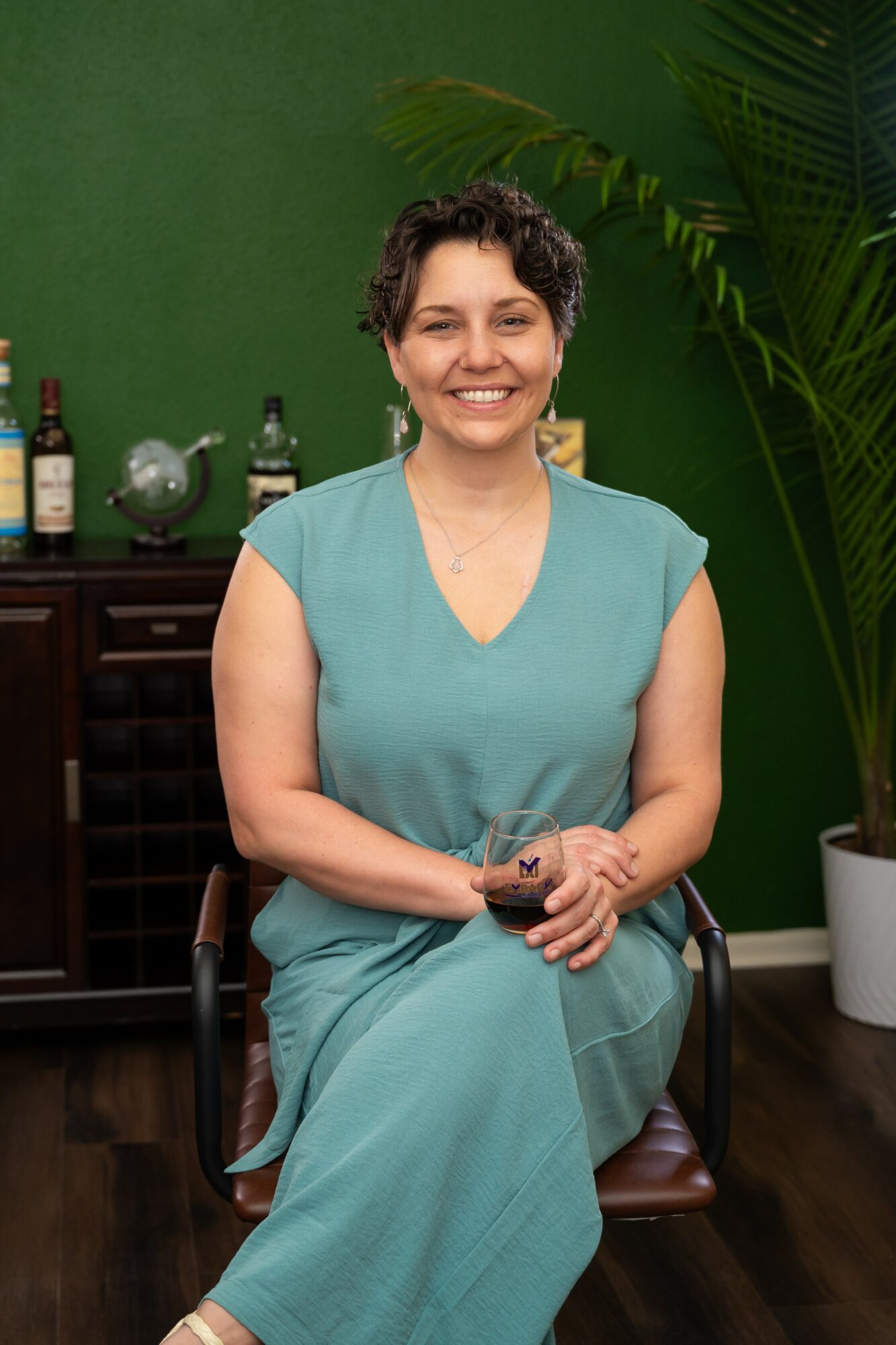Smiling woman with short curly hair in teal dress sitting on a chair in front of a green wall and large plant.
