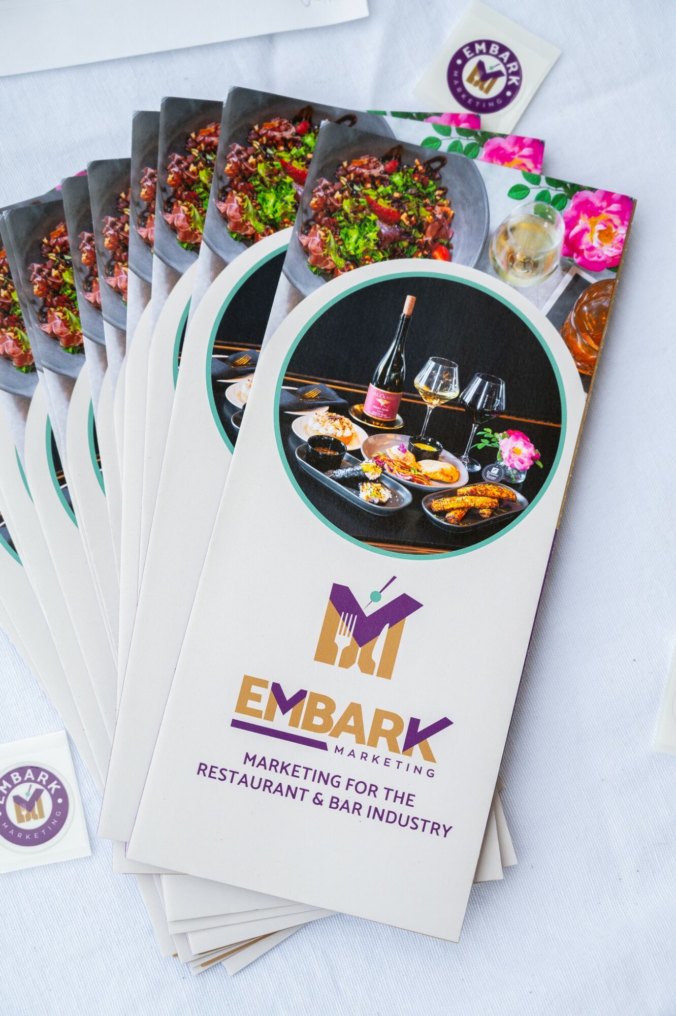 Brochure titled 'EMBARK' with a circular image of a sushi platter and a wine bottle, and colorful flowers, for restaurant and bar industry marketing.