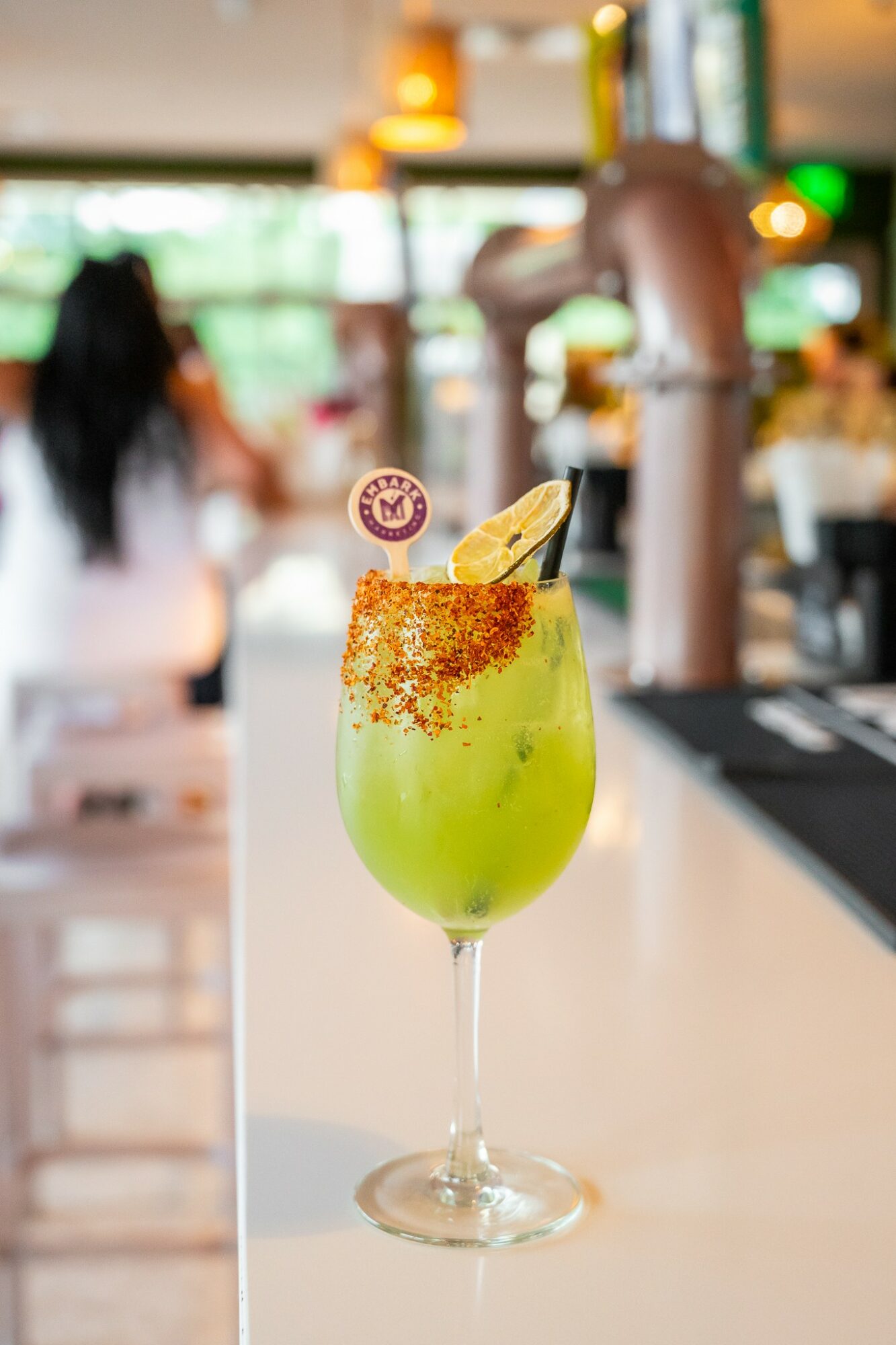 Green cocktail with a lemon wedge and a cherry garnish in a tall glass, blurred background of a cafe or bar, 20 words