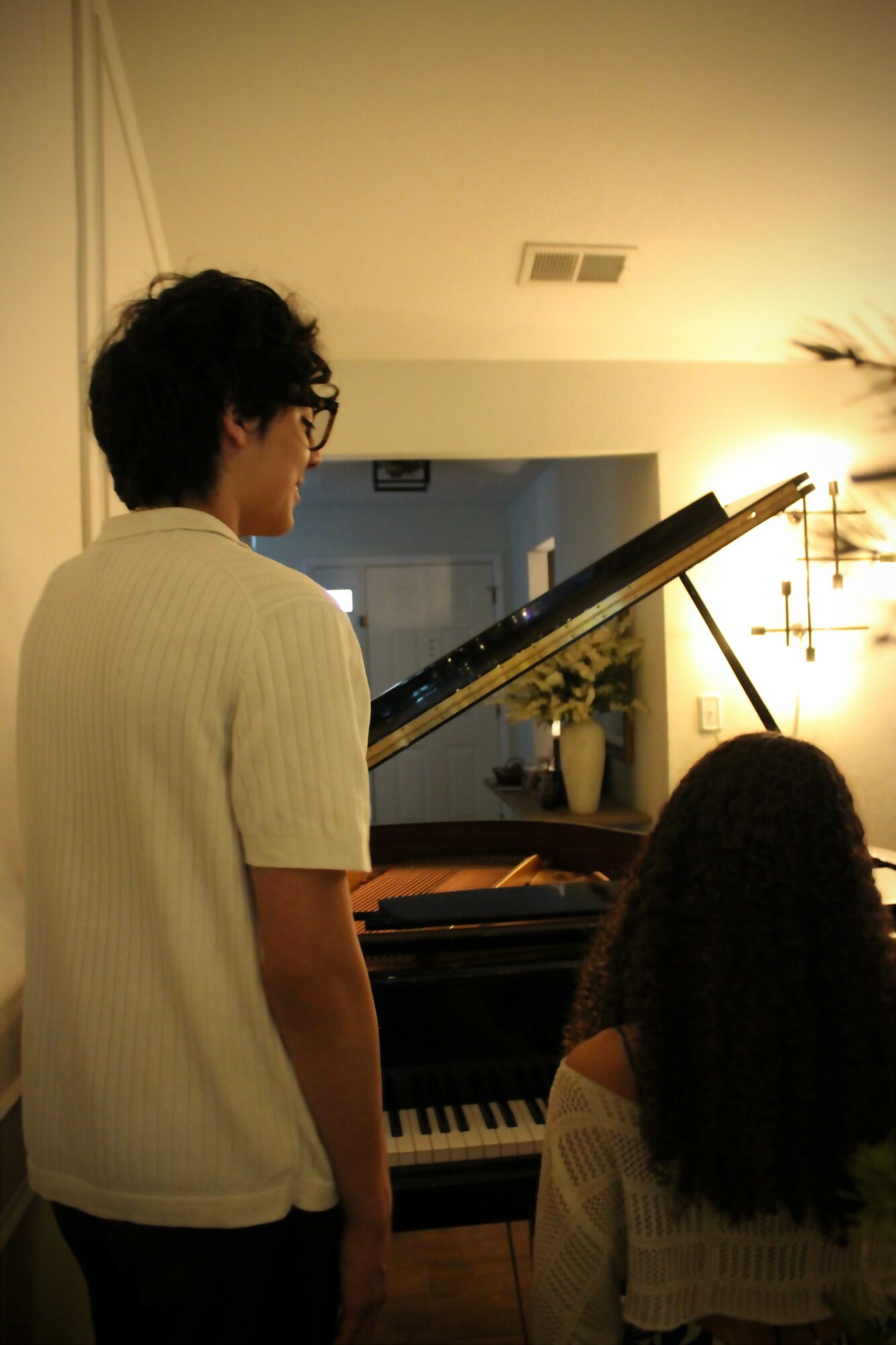 Person with glasses and short hair stands near a piano, looking at sheet music, in a warmly lit room.