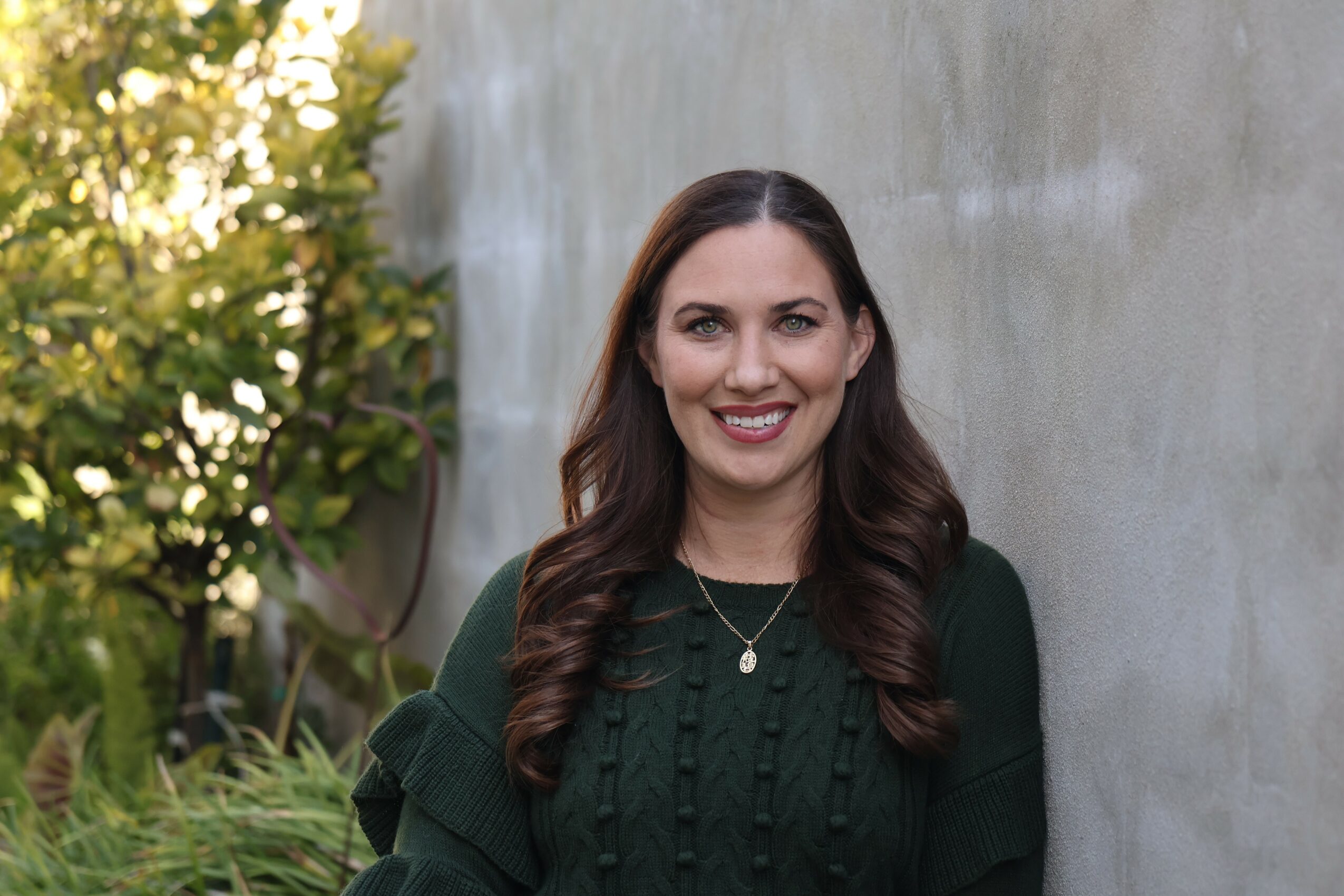 Smiling woman with long dark hair wearing a dark green sweater outdoors near a concrete wall.