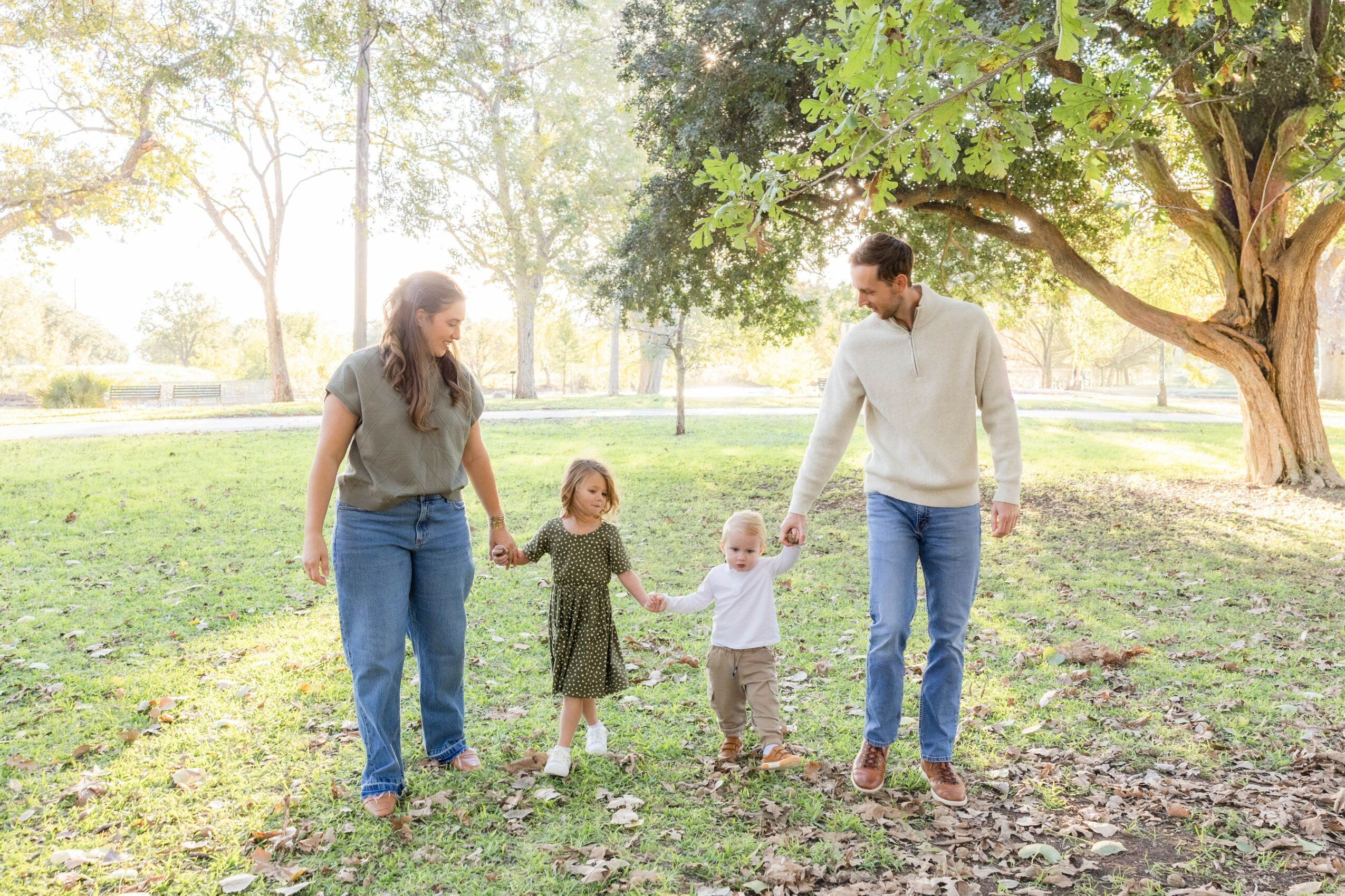 Family of four walking in a park holding hands, trees and sunlight in the background.