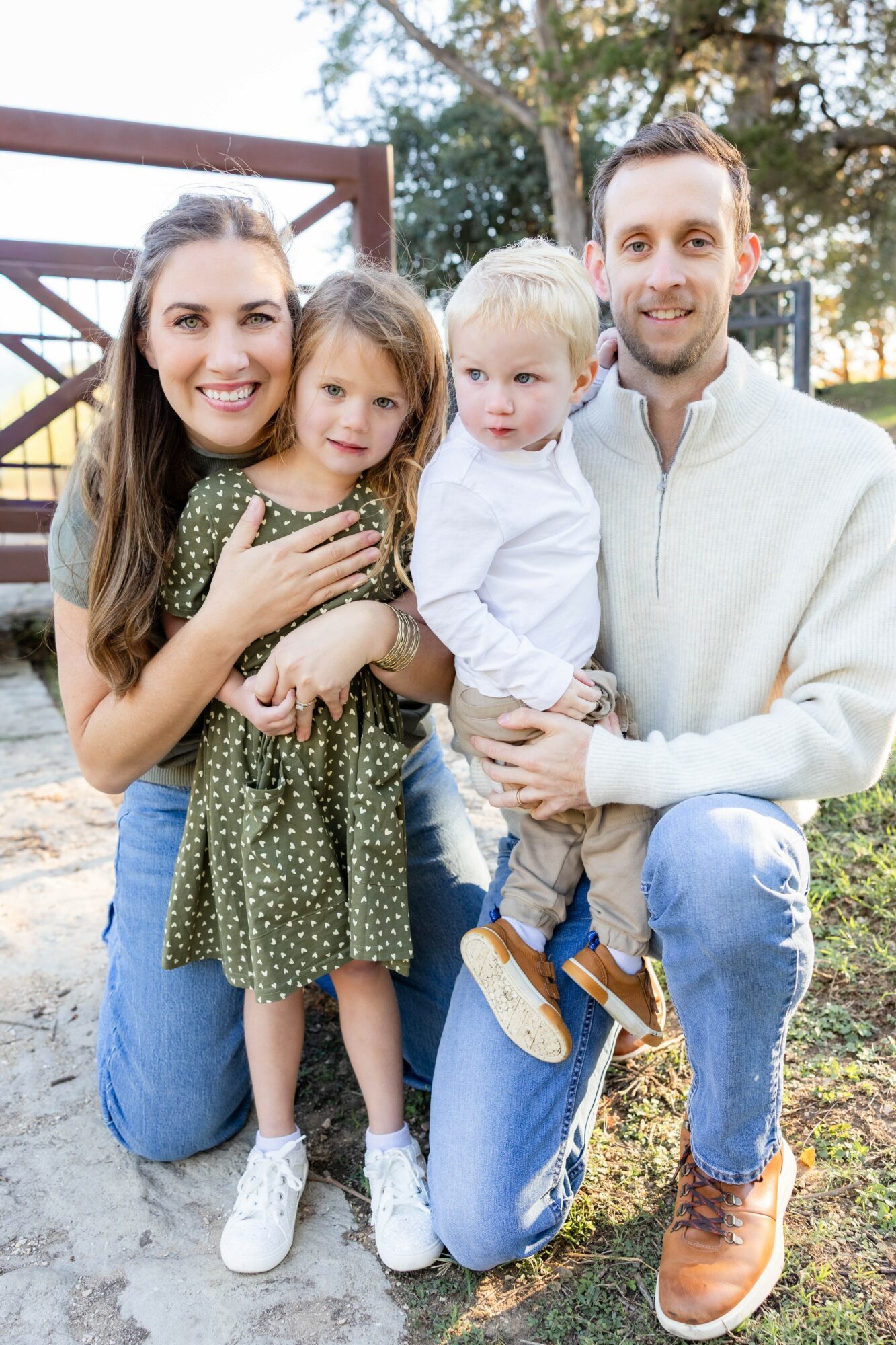 Family of four outdoors, smiling, with trees and playground equipment in background.