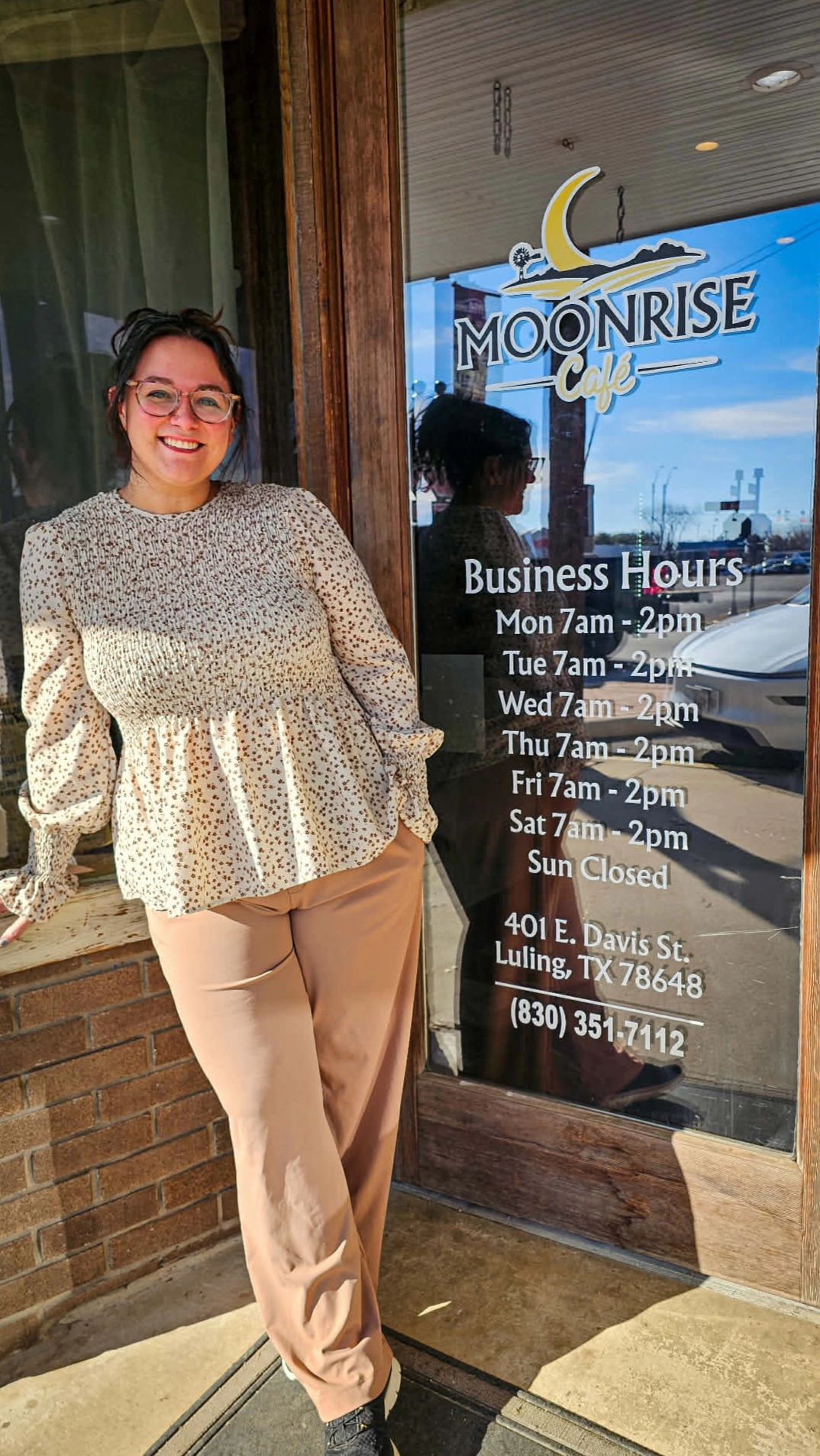 Woman with glasses smiling standing outside a business with moon and sunrise sign, reflection in window, casual clothing.