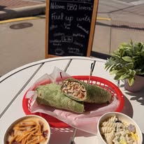 A burrito on a red tray with a side of pasta and a small bowl of salad, on a white table outdoors.