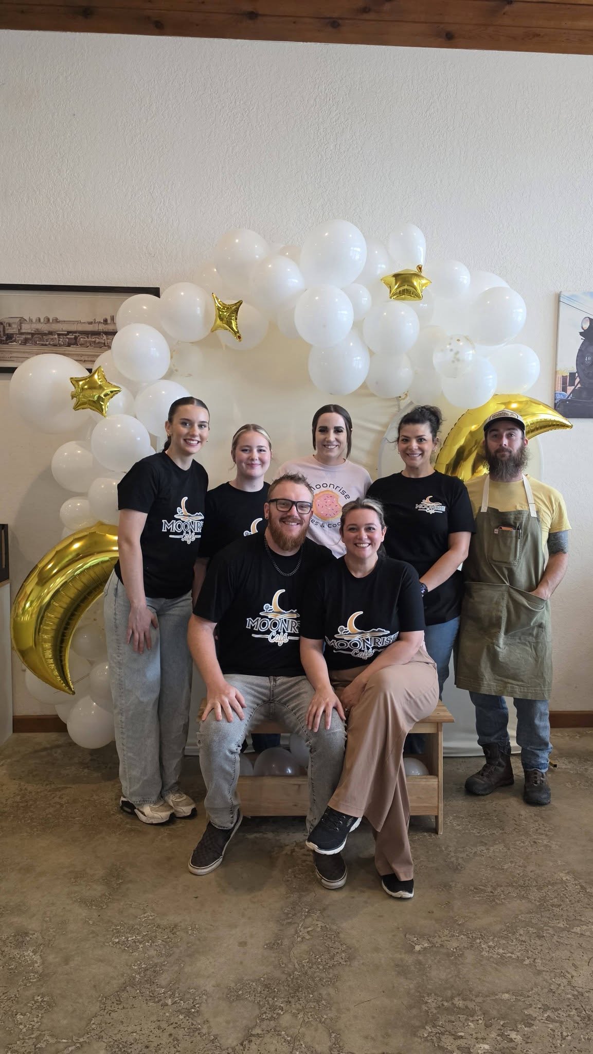 Group of six people posing in front of balloon decorations with white, gold, and star-shaped balloons, indoors.