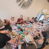 Children and adults sit around a table with food and drinks, engaging in a meal or celebration indoors.