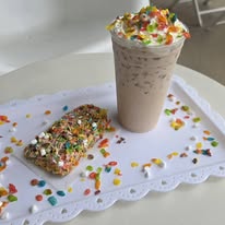 A milkshake topped with whipped cream and colorful sprinkles, alongside a decorated rice crispy treat on a white tray.