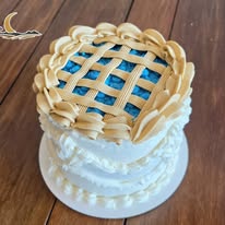 Round cake with white frosting, decorative cream border, and a blue lattice top, on a wooden surface.