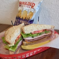 Sandwich with lettuce, tomato, ham, and cheese on bread, with a pickle, in a red basket, and a bag of potato chips behind.