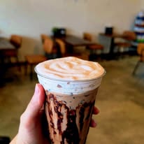 Hand holding a caramel frappuccino with whipped cream in a clear plastic cup, inside a cafe with tables and chairs.