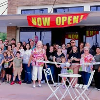 Group of people gathered outside a store with a 'Now Open' banner, celebrating opening day, with a woman and child cutting a ribbon.