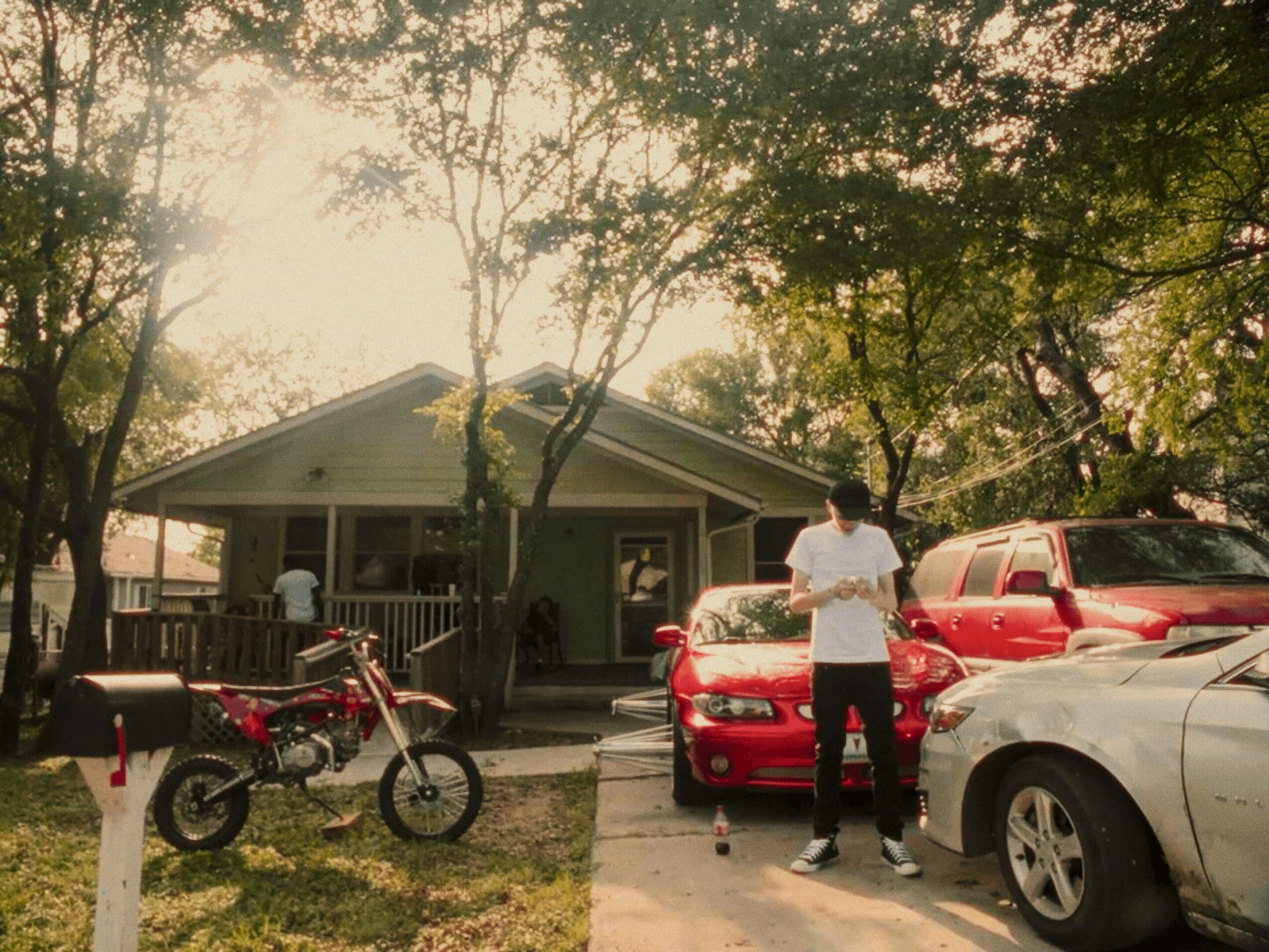 Person standing in front of a red car, with a motorcycle and parked vehicles nearby, in front of a house surrounded by trees.