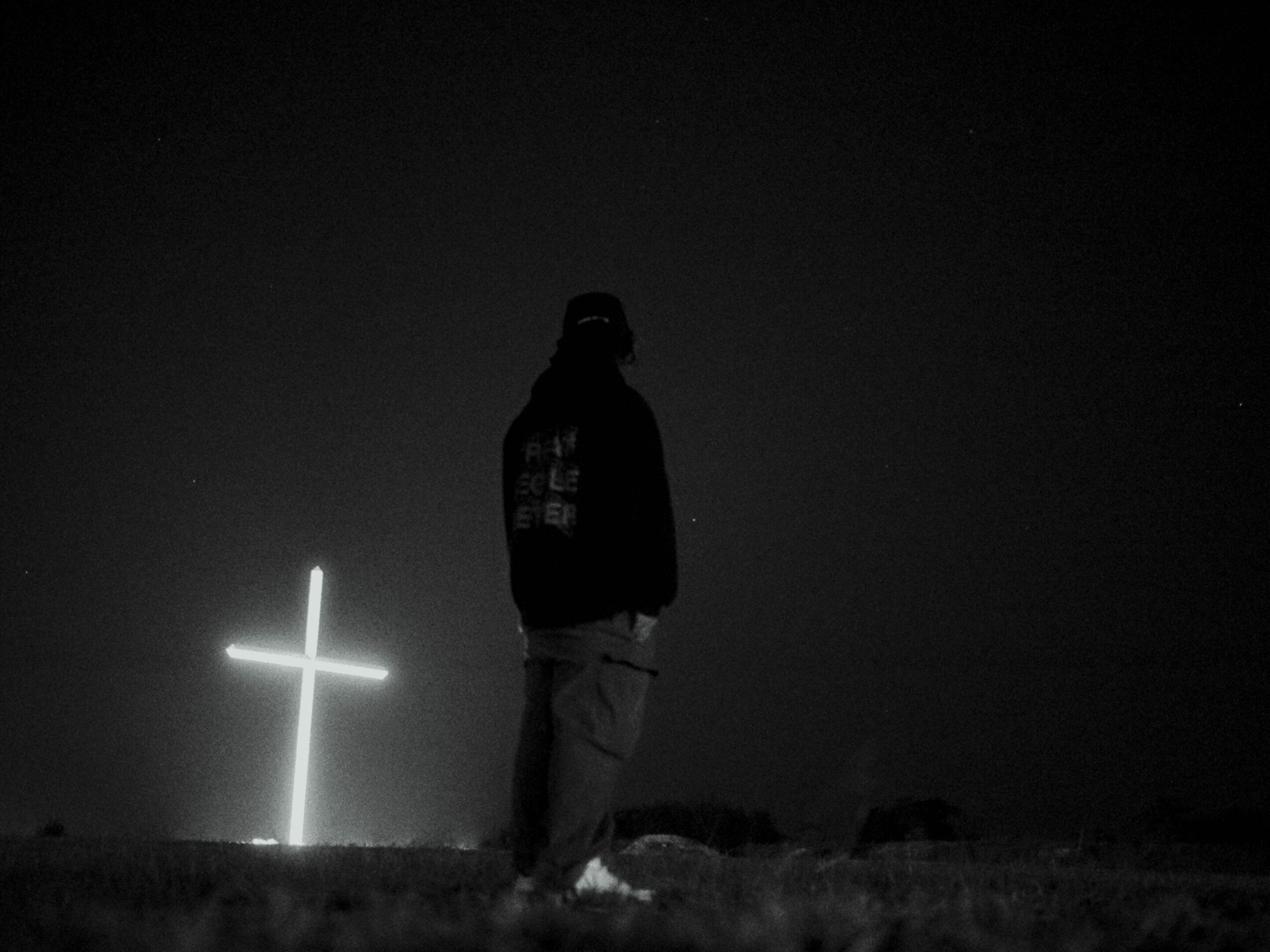 Person standing outdoors at night near a glowing cross, dark sky, and grassy ground, in black and white.