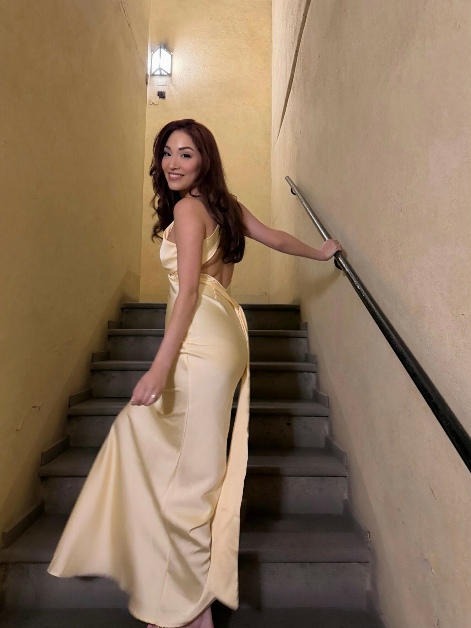 Young woman in a cream-colored gown standing on stairs, holding railing, smiling, with warm lighting above.