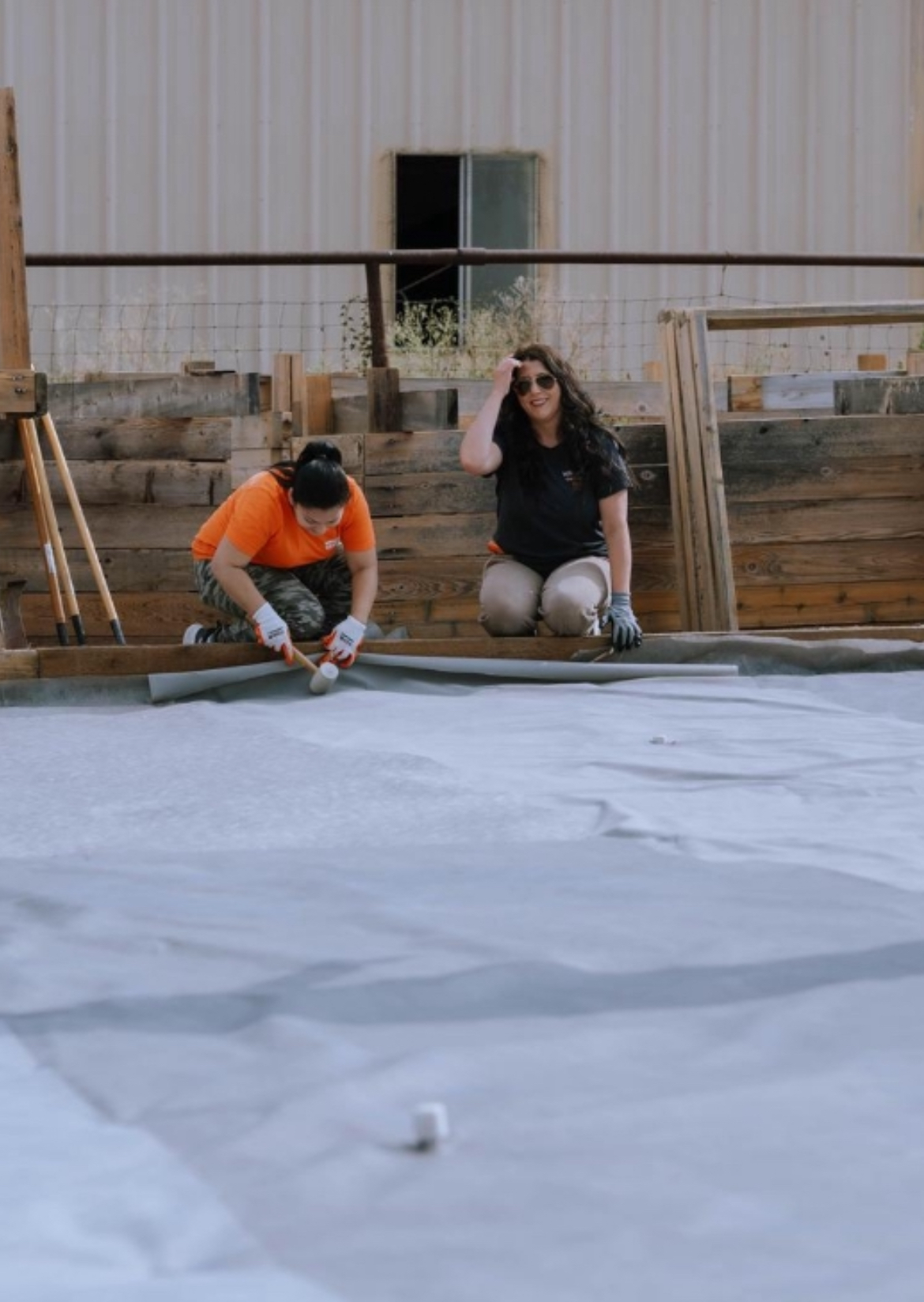 Two people kneeling on a surface, one in an orange shirt and the other in black, with a building in the background.