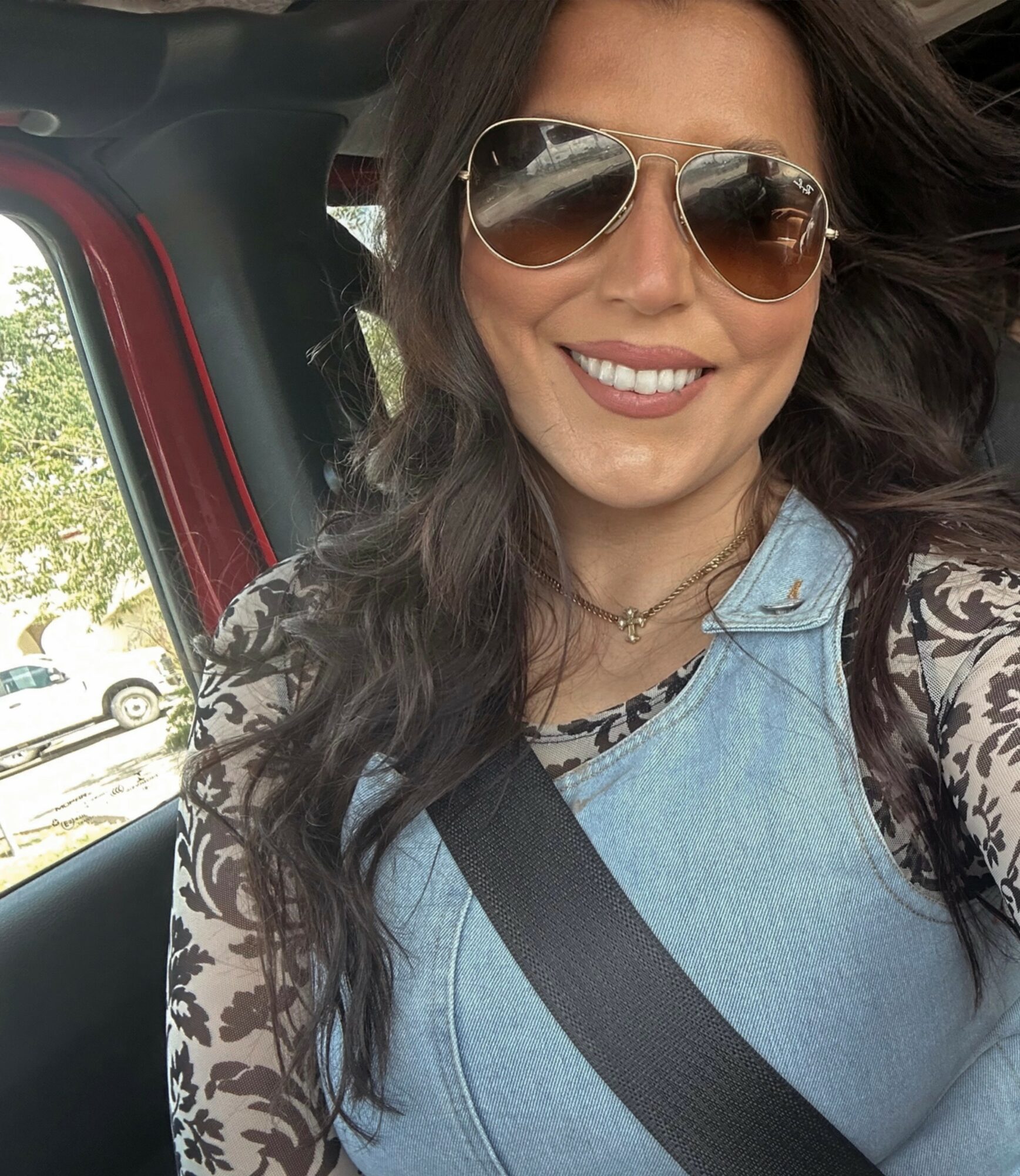 Woman with long dark hair wearing sunglasses, a denim top, and a patterned long-sleeve shirt, smiling inside a vehicle.