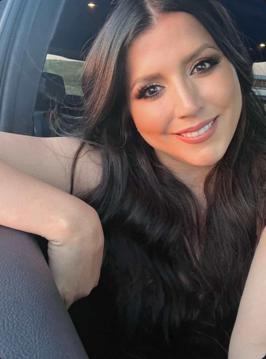 Smiling woman with long dark hair sitting in a car, arm resting on the seat, natural light, casual setting.