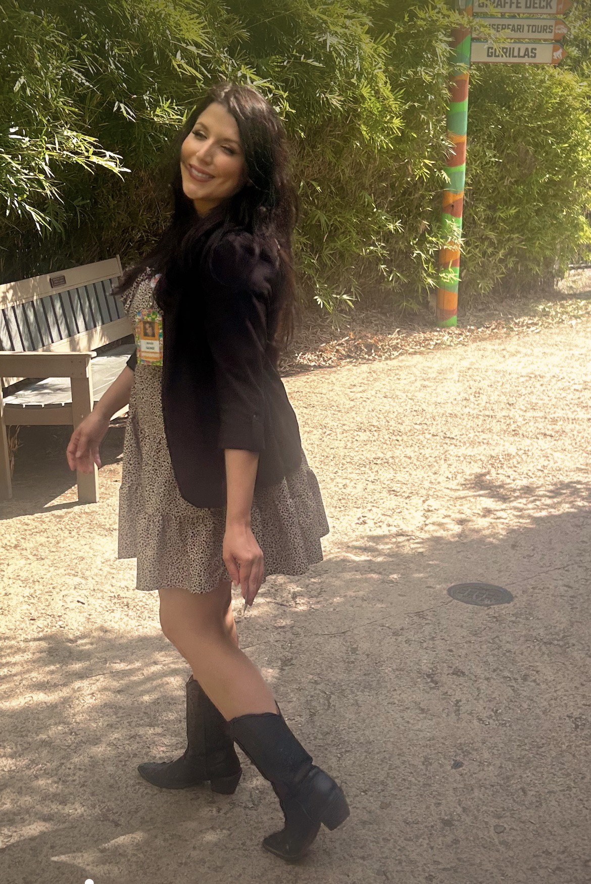 Young woman with long dark hair smiling outdoors, wearing a black jacket, patterned dress, and black boots, standing on dirt ground.