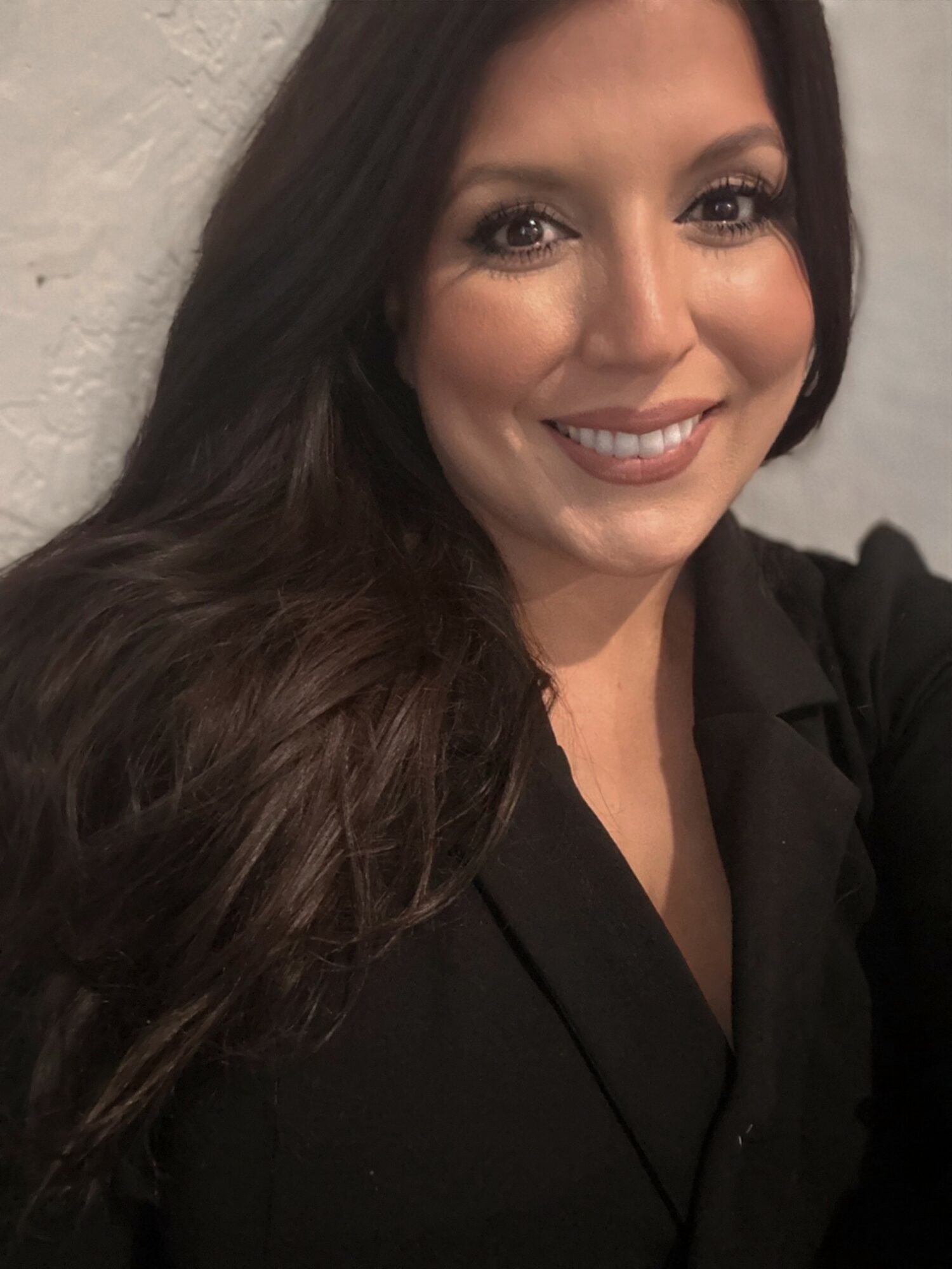 Smiling woman with long dark hair wearing a black top, standing against a light-colored wall.