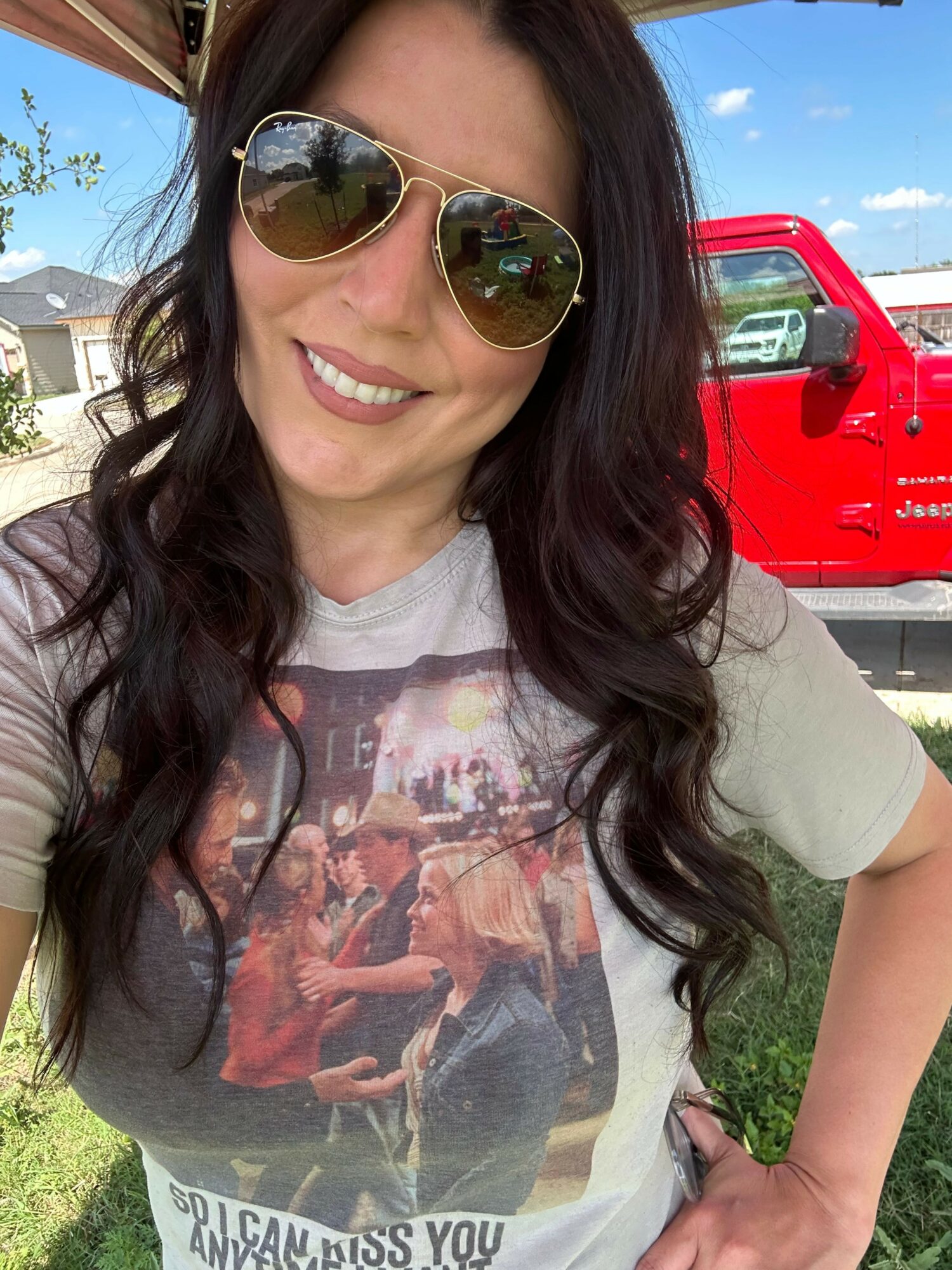 Woman with long dark hair wearing sunglasses and a graphic t-shirt, smiling outdoors with a red truck in background.