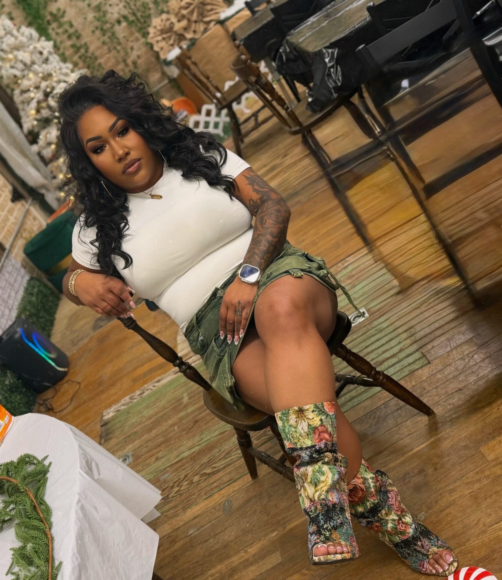 Woman with curly hair and tattoos sitting on a wooden chair, wearing a white top, camouflage shorts, and floral leg warmers, in a room with wooden floors and furniture.